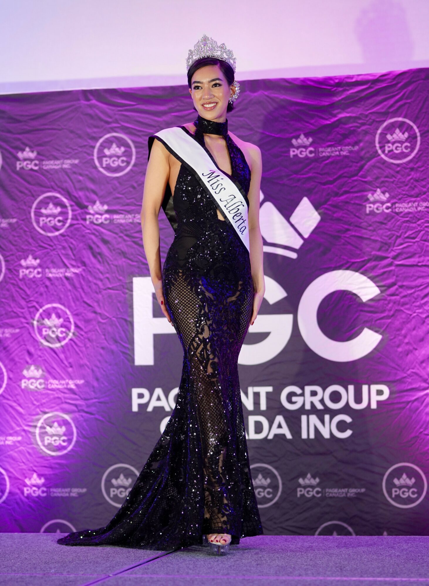 Woman wearing a black sequined gown and crown, holding a sash, standing on stage with a purple backdrop.