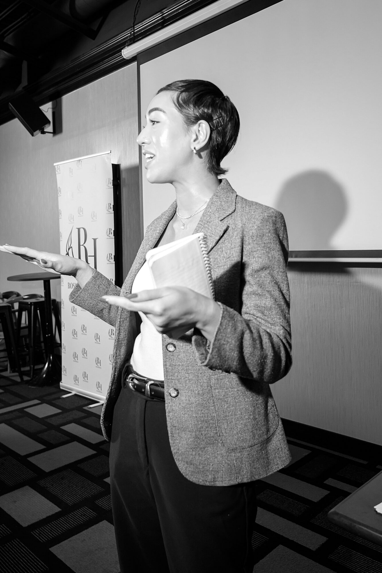Woman in blazer holding papers, smiling, standing indoors with a projection screen behind her.