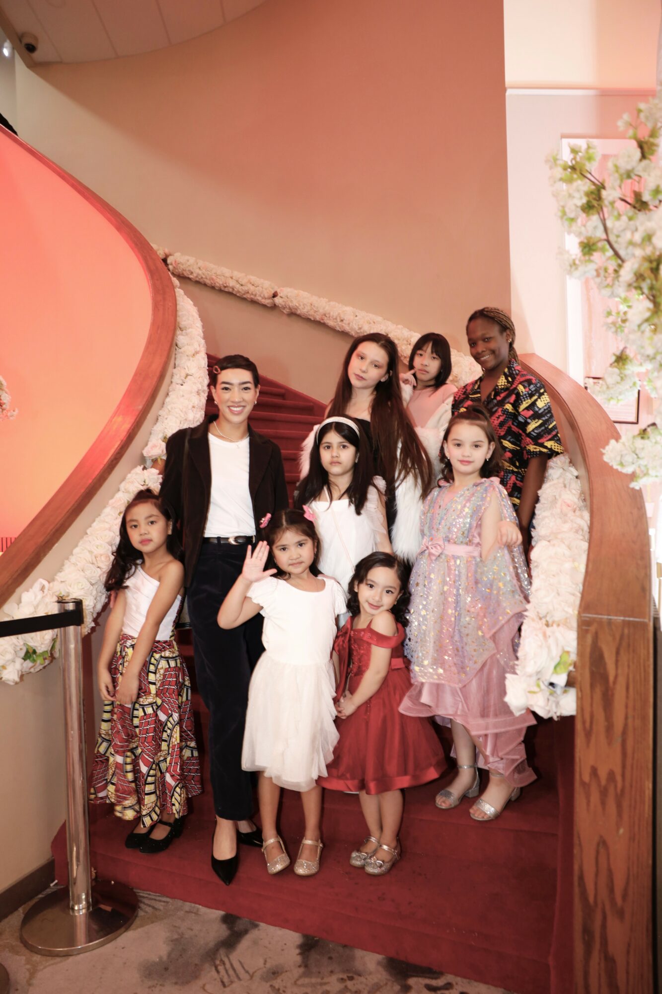 Group of nine women and girls posing on a staircase with pink walls and floral decorations, smiling at the camera.