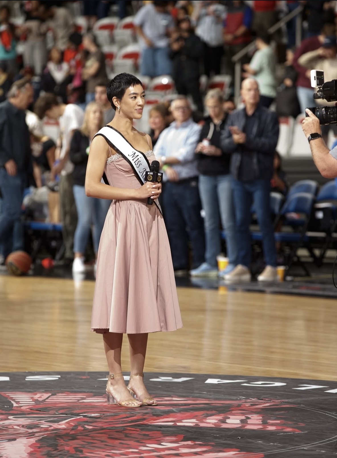Young woman in a pink skirt and sash stands on a basketball court, holding a trophy, with a crowd and camera in background.