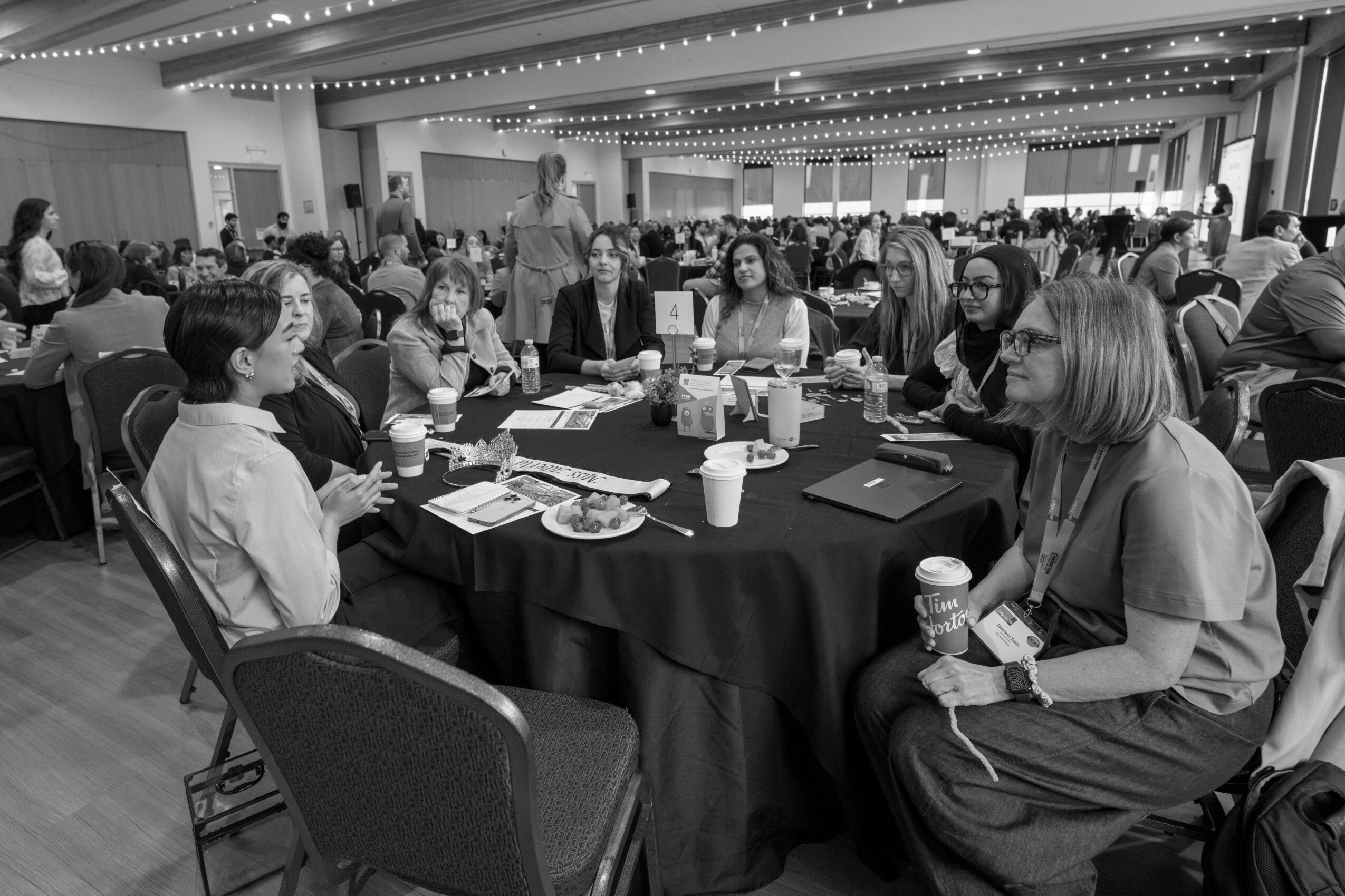 Group of people sitting around a round table in a large conference room, engaged in discussion, with food and drinks on the table.