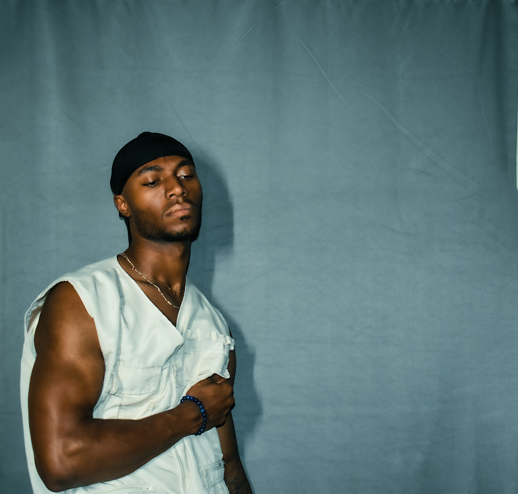 Young man with a black head covering, wearing a sleeveless white shirt, standing against a plain background.