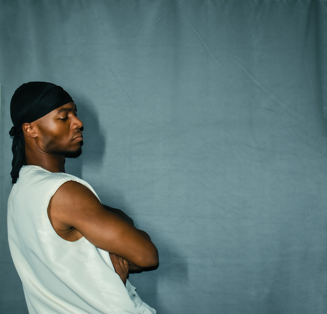 Side view of a person with arms crossed, wearing a black head covering and white sleeveless shirt, against a plain background.