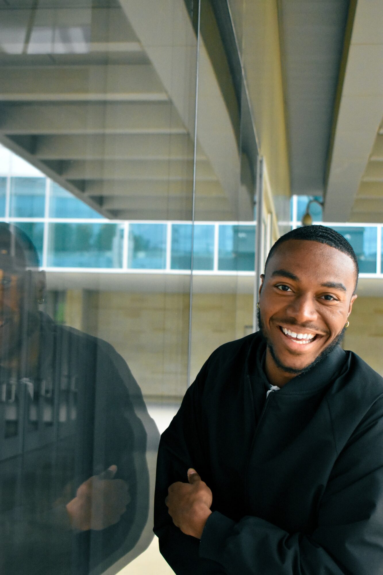 Smiling man in black jacket standing near glass window with modern building background.
