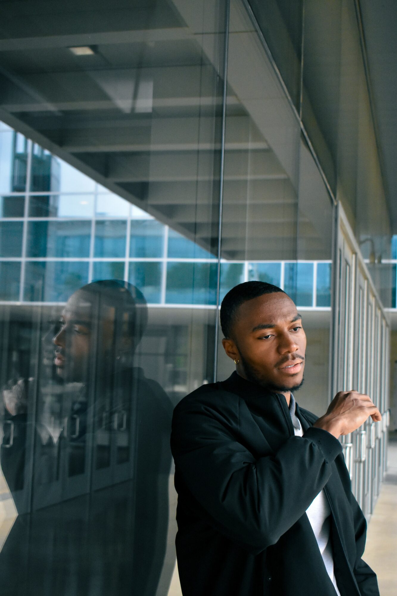 Man with short hair and beard in black jacket stands near glass wall, reflection visible, modern building background.