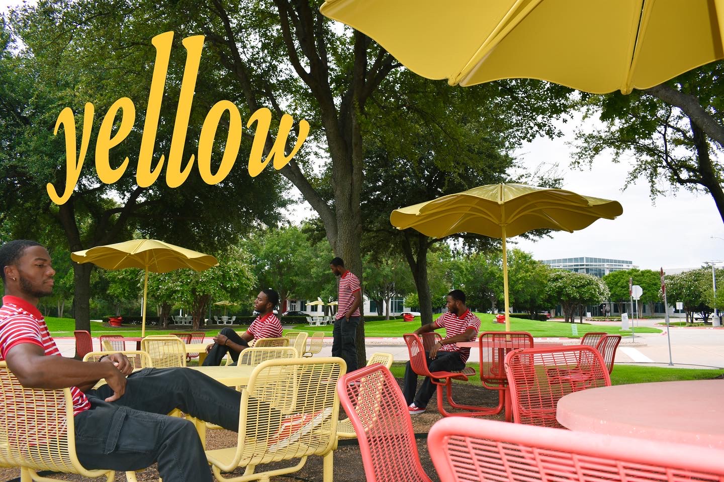People sitting at outdoor tables with yellow umbrellas in a park, trees and grass in background, sunny day.
