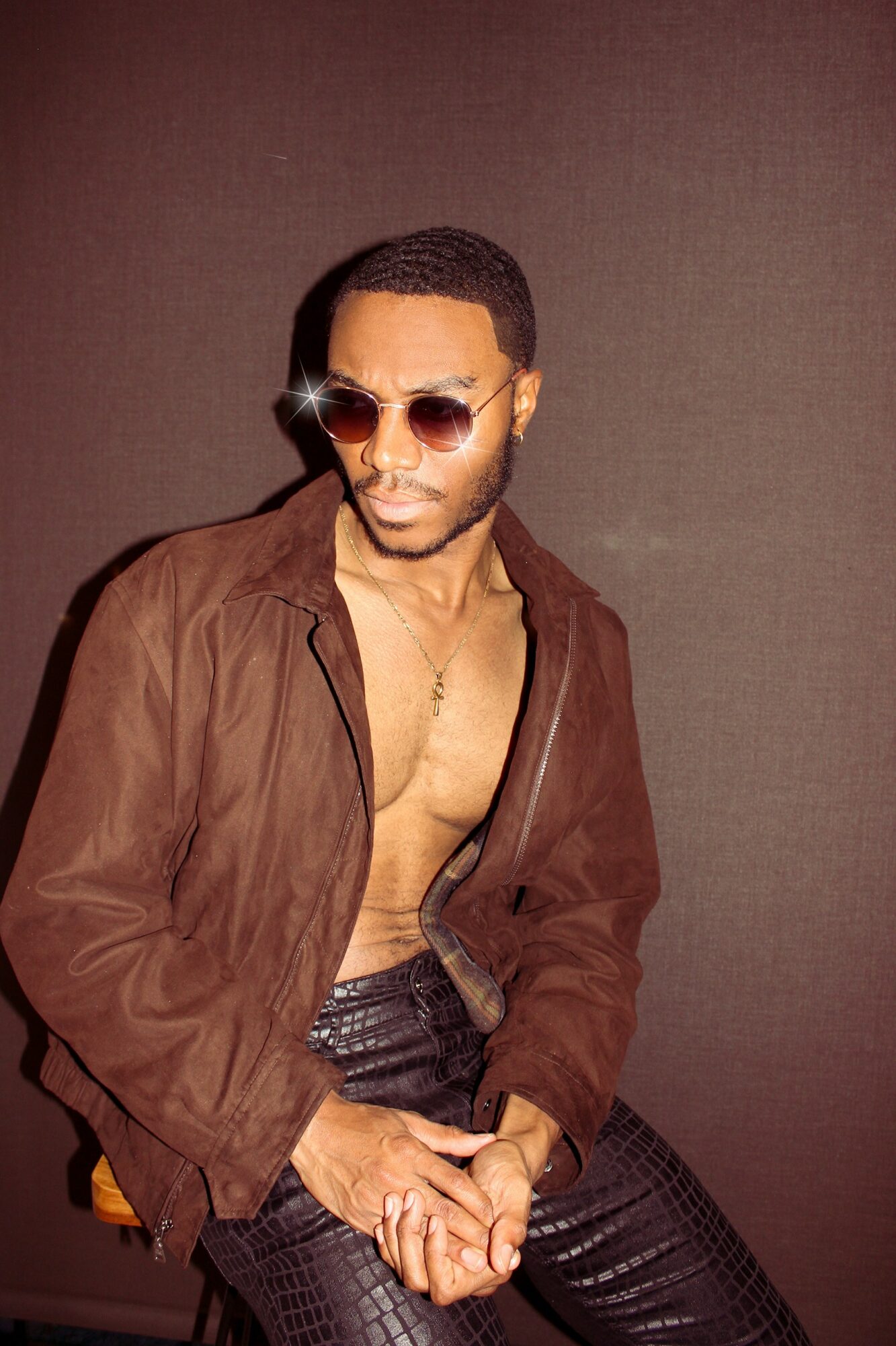Man with sunglasses wearing a brown jacket and black pants, posing against a plain background.