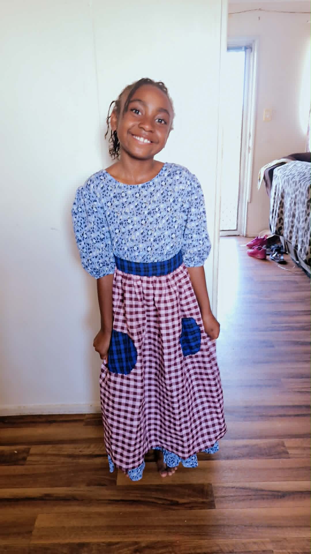 Young girl smiling, wearing a blue patterned top and a long checkered skirt with pockets, standing indoors.