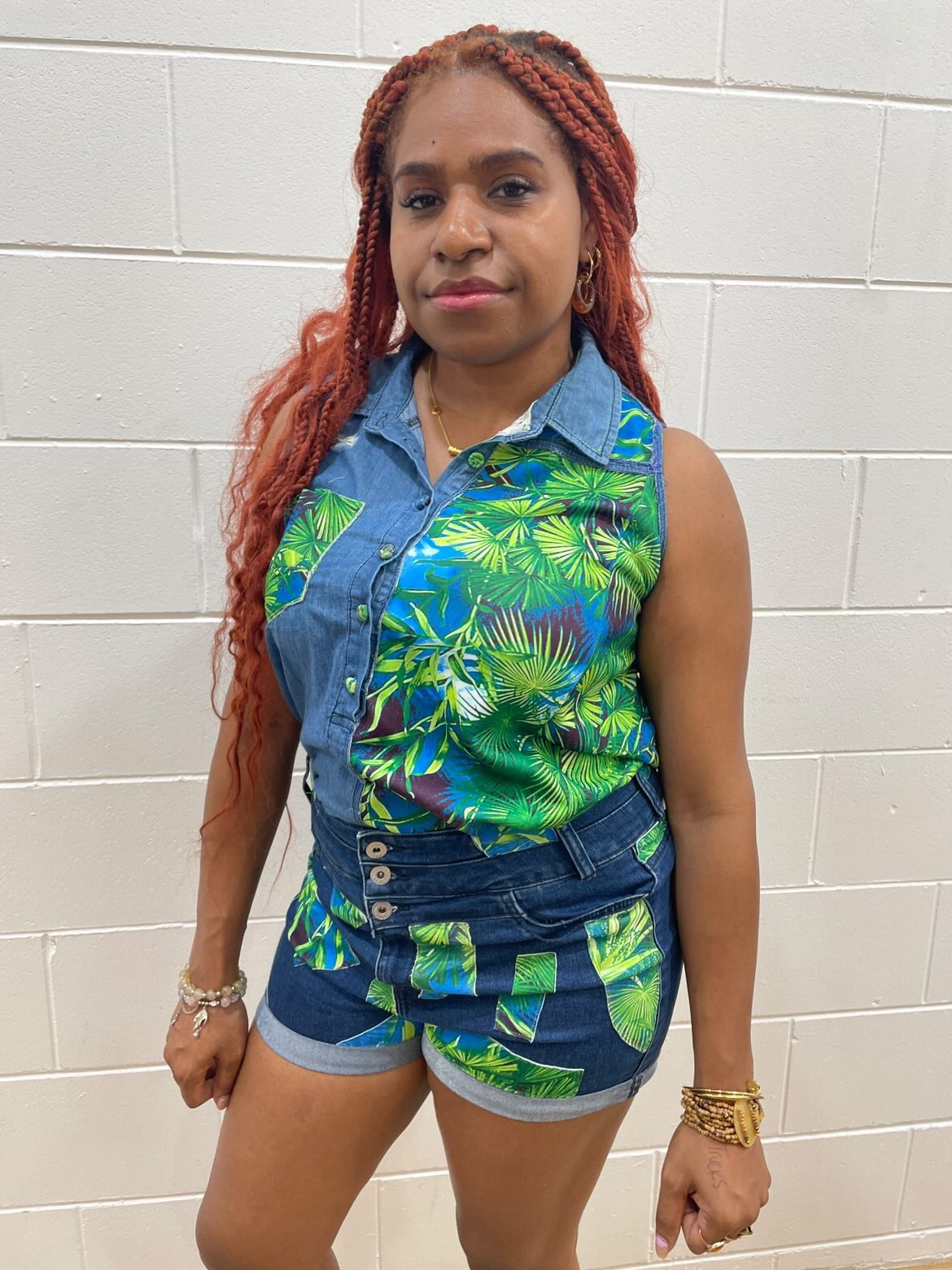 Young woman with long red hair wearing a sleeveless tropical print top and denim shorts, standing against a white tiled wall.