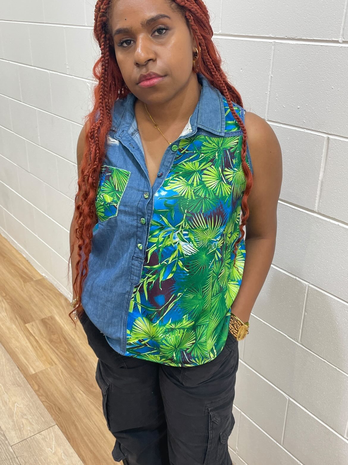 Young woman with long red braided hair stands against a white tiled wall, wearing a colorful tropical sleeveless shirt and black pants.