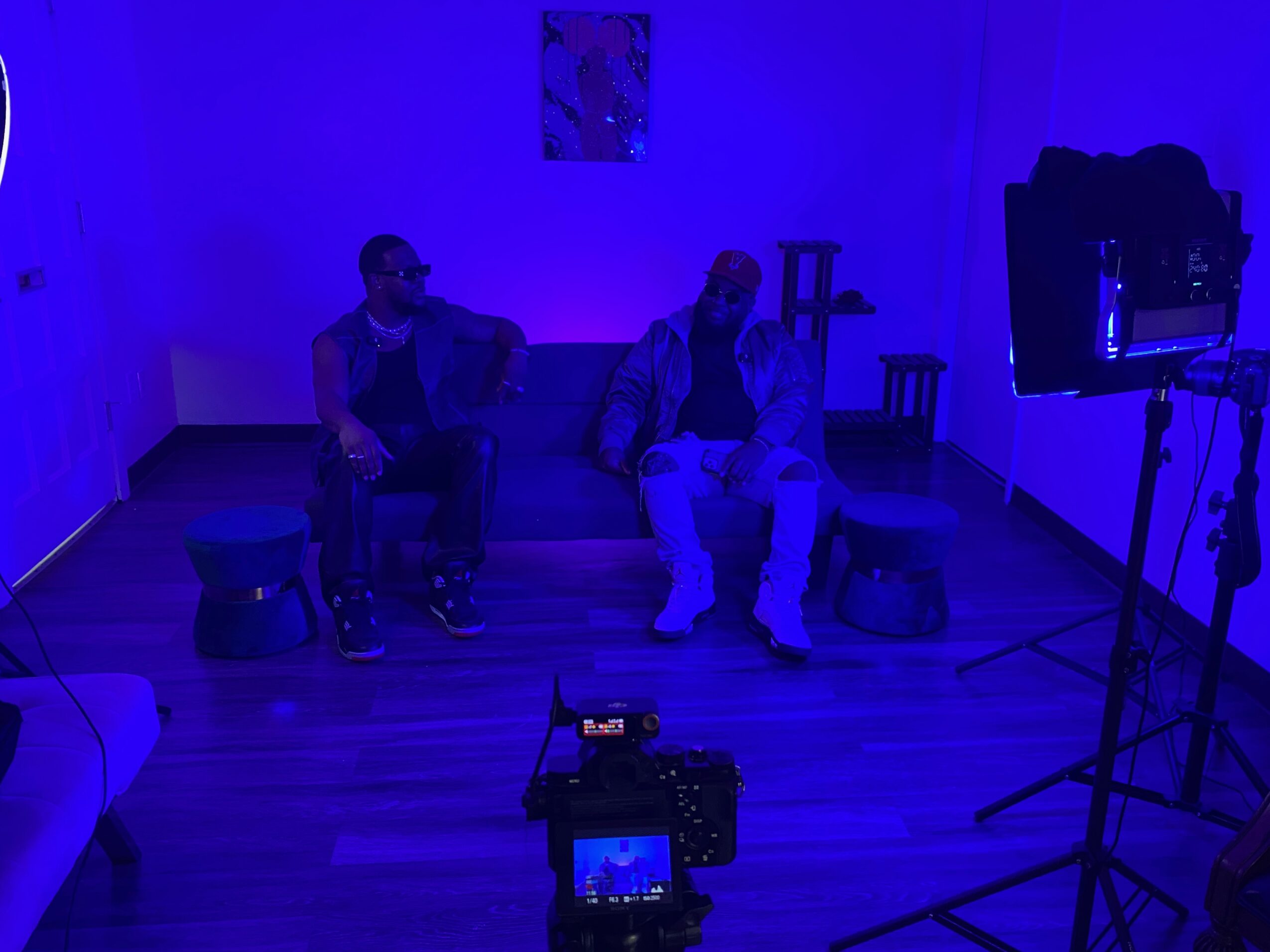 Two people sit on a sofa in a dimly lit room with blue lighting, one with a dog nearby, and a camera on a tripod.