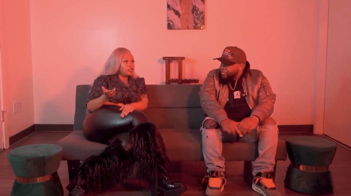 Two people sit on a sofa in a room with pink lighting, facing each other, with small tables nearby.