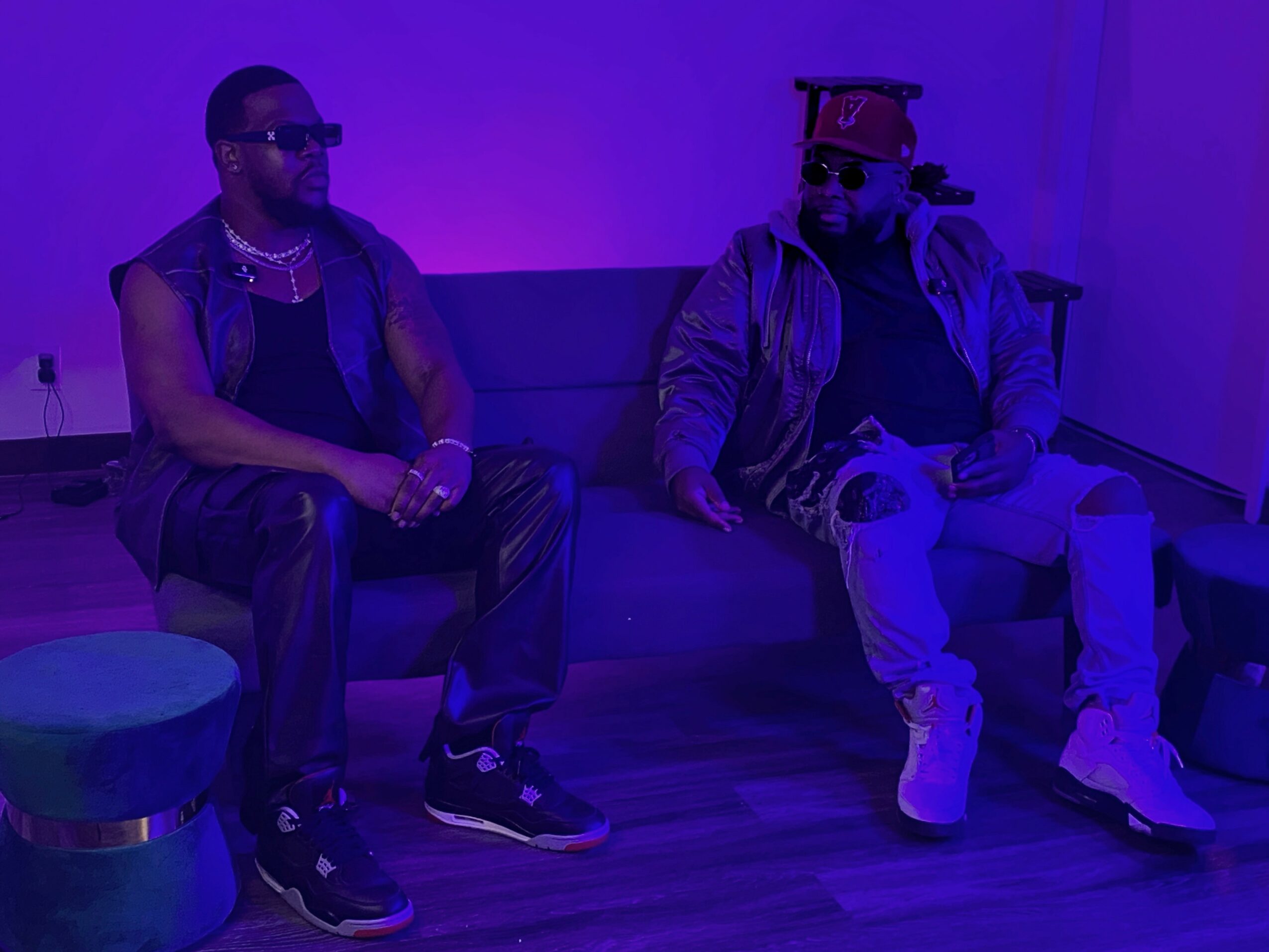 Two men sit on a sofa in a room with purple lighting, wearing sunglasses and casual clothing.