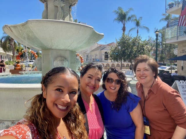 Four women smiling in front of a fountain outdoors on a sunny day, with trees and buildings in the background.