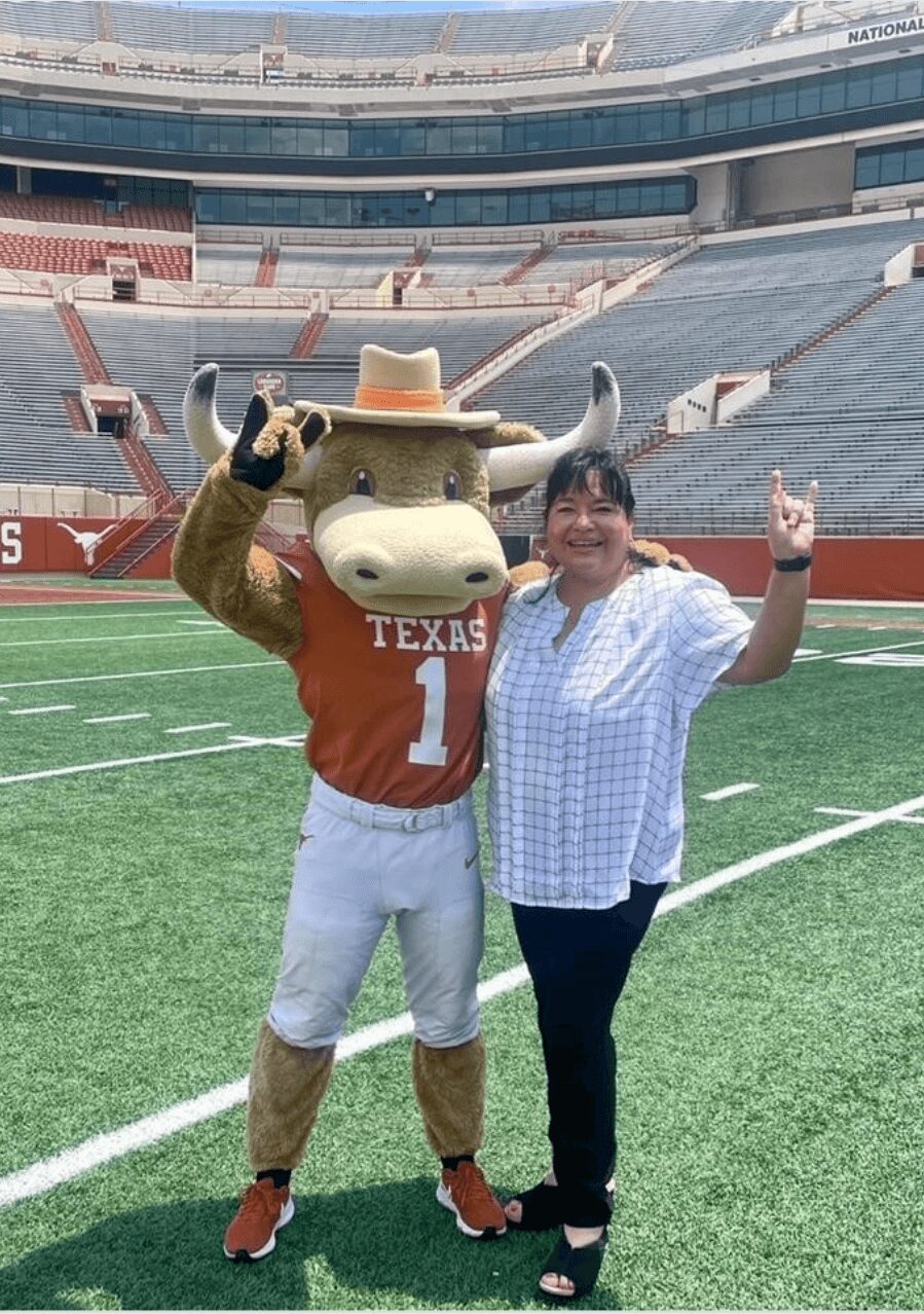 Person standing next to a Texas Longhorns mascot on a football field, both smiling and making a peace sign.