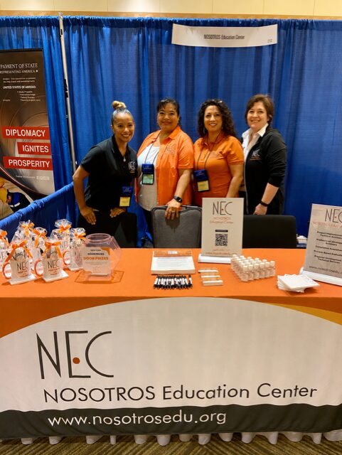 Four women stand behind a table at an educational event, with a blue curtain backdrop and NEC signage, smiling at the camera.