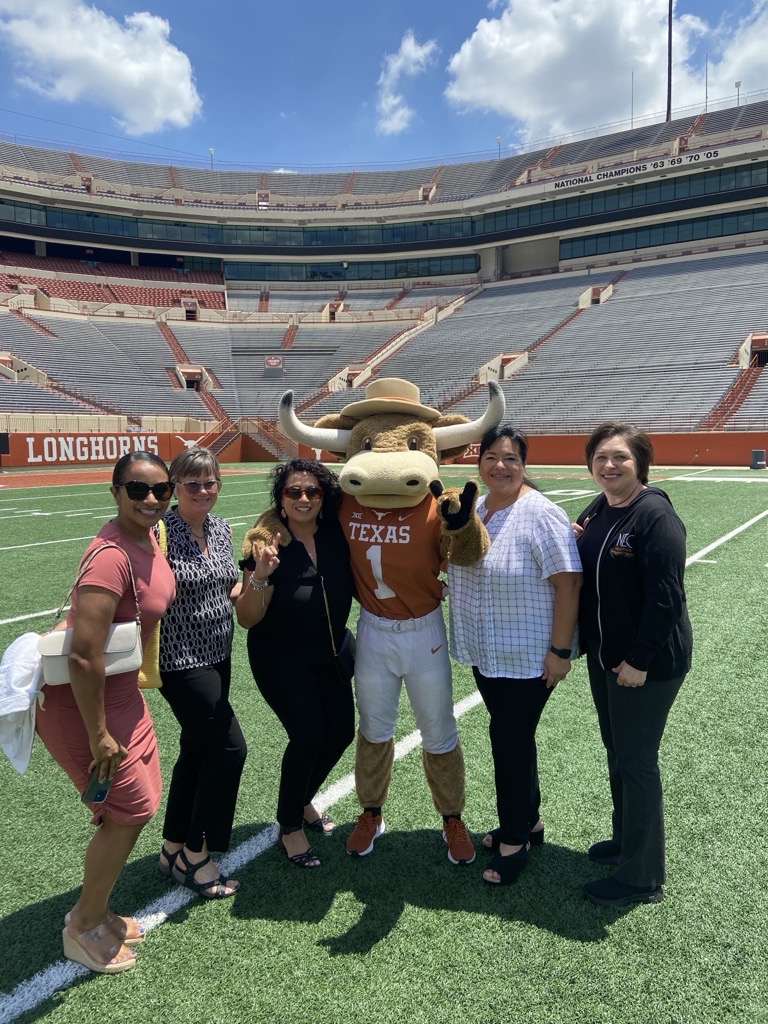 Group of five women and a mascot on a football field in a stadium, smiling and posing for a photo.