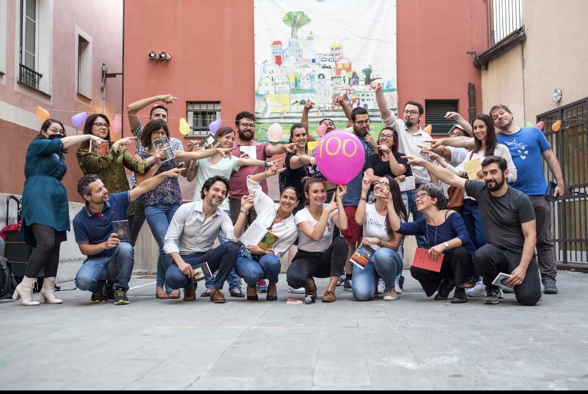Group of people outdoors, some holding a pink balloon, posing with arms extended towards the balloon, in front of a colorful mural.