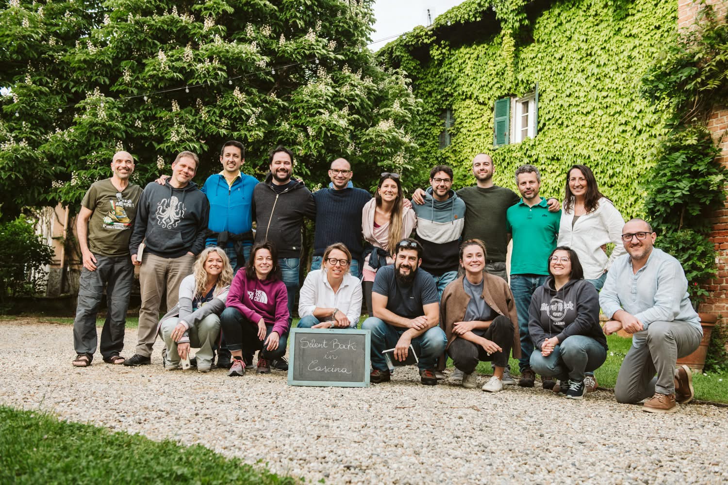 Group of 15 people outdoors, some standing and some kneeling, with greenery and a building in the background.