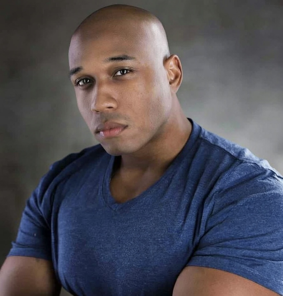 Portrait of a young man with a shaved head wearing a blue shirt, looking at the camera with a neutral expression.