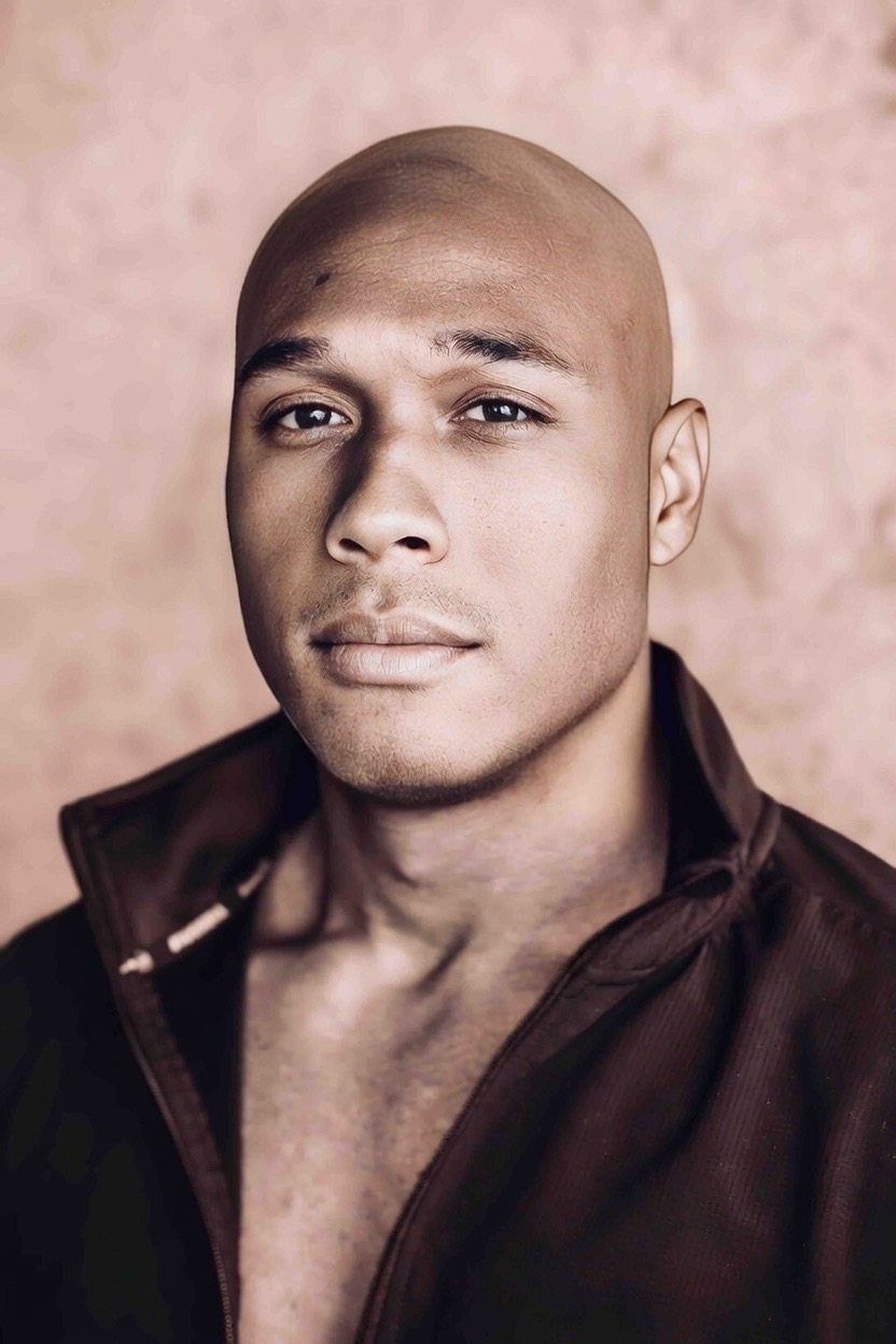 Portrait of a bald man with a serious expression, wearing a dark jacket, against a pinkish background.
