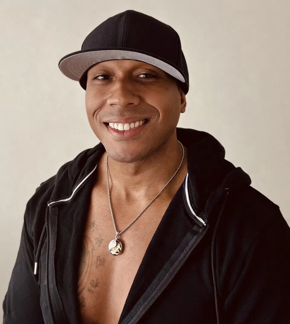 Smiling man wearing a black cap, black hoodie, and a necklace, with a light background.