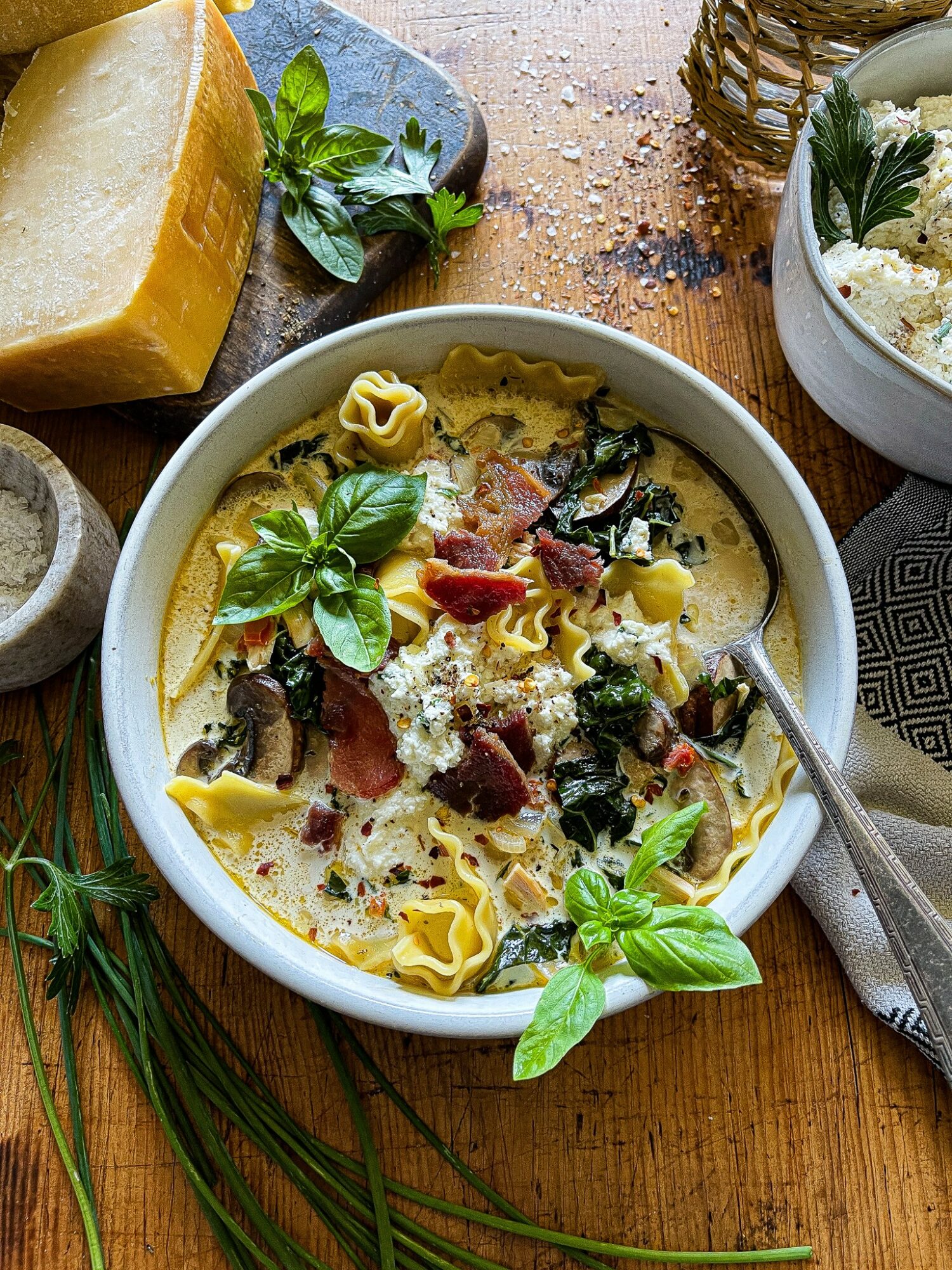 Bowl of pasta with herbs, cheese, and dark toppings on a wooden table, surrounded by lemon, herbs, and a candle.