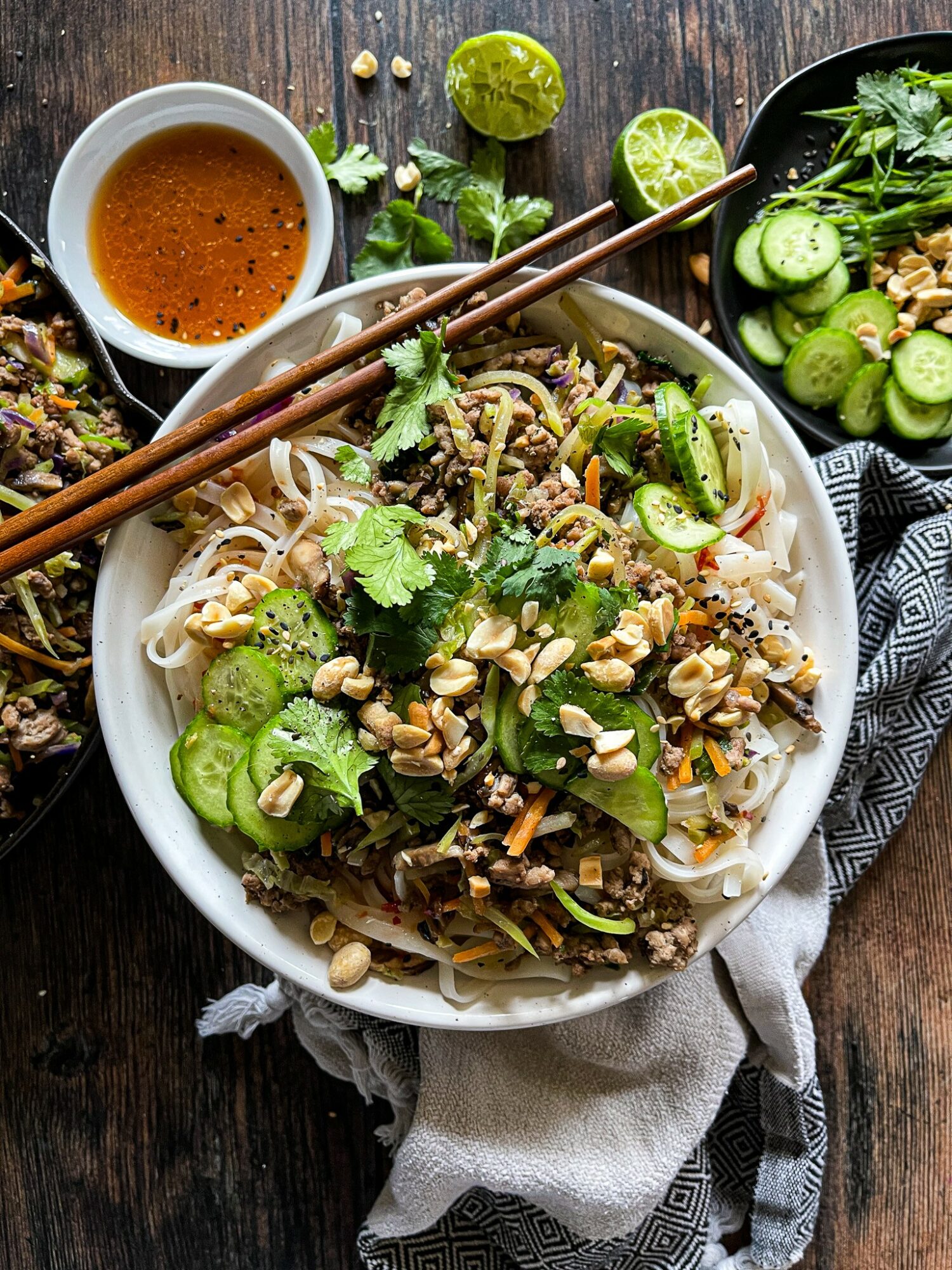 Bowl of noodle salad with cucumbers, peanuts, cilantro, and lime slices on a wooden table, with dipping sauce and lime nearby.