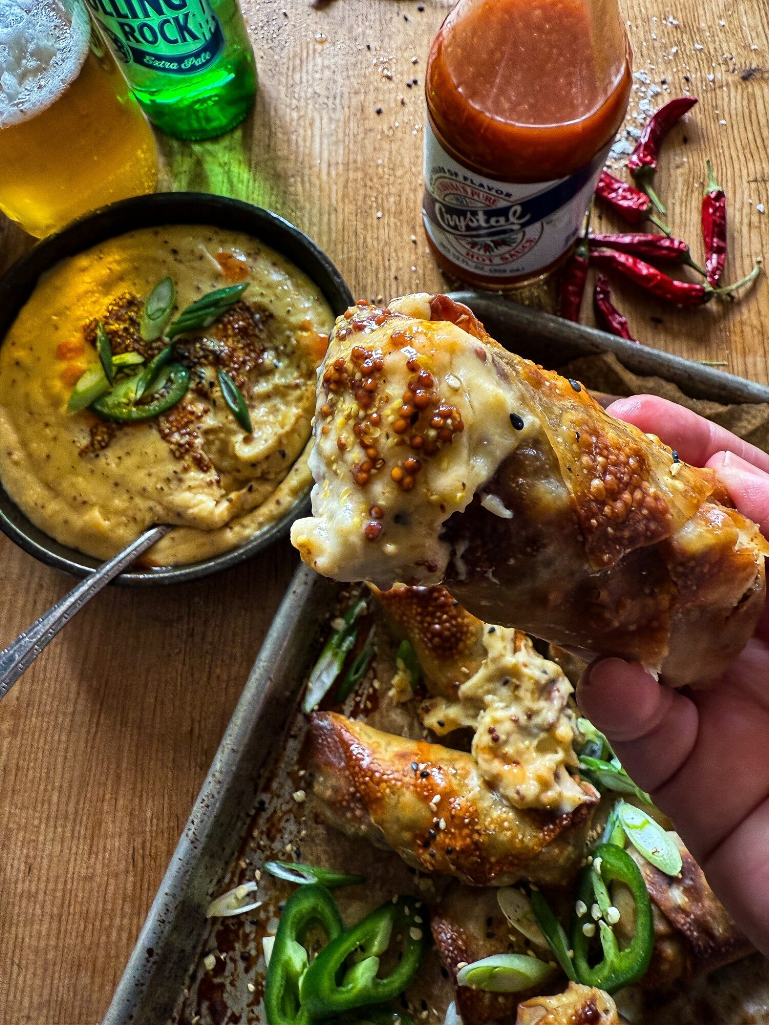 Person holding a slice of pizza with cheese, sauce, and toppings, with a bowl of dip, hot sauce, and drinks on table