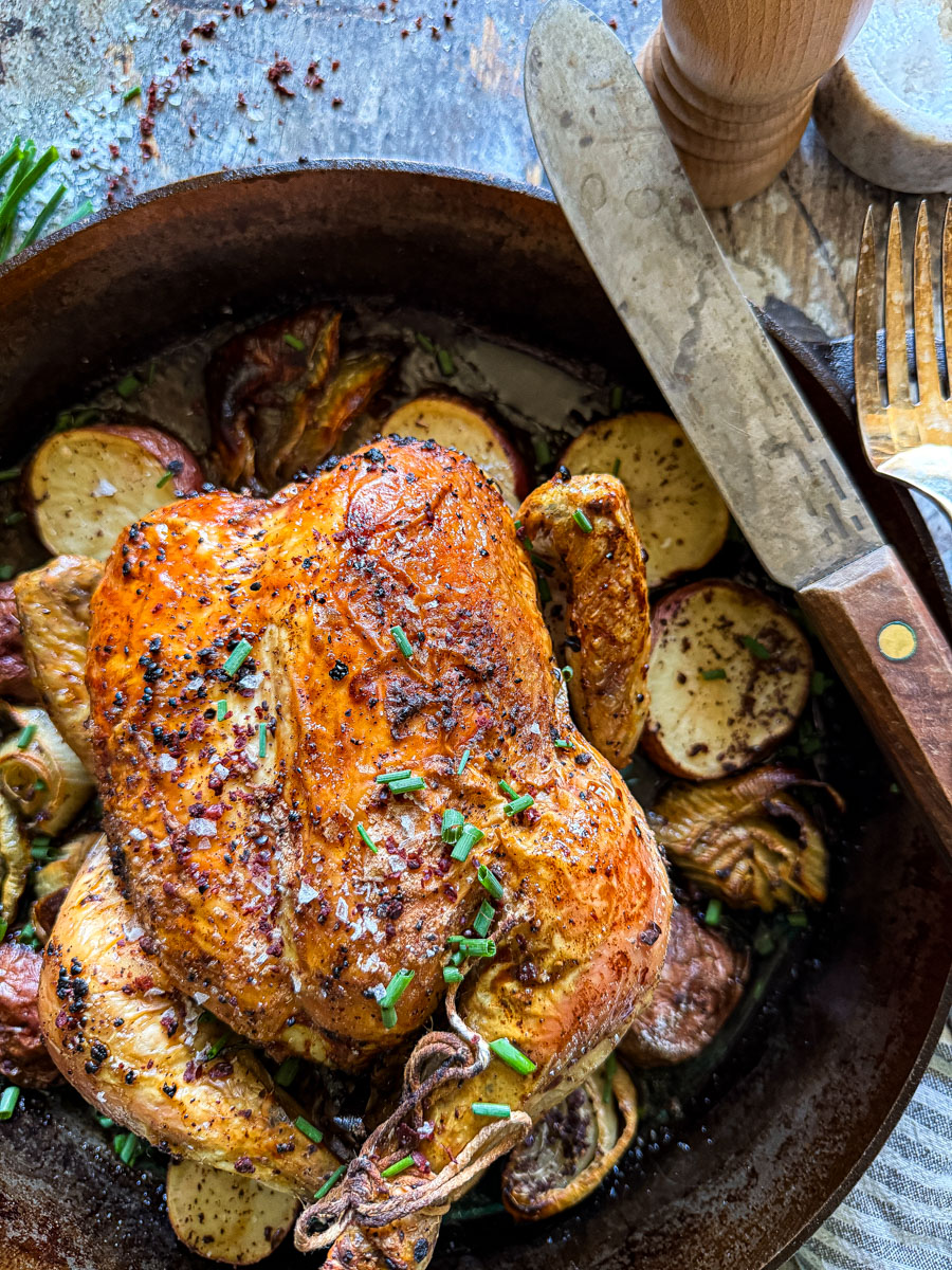Roasted chicken with herbs and vegetables in a skillet, garnished with chopped herbs and colorful seasoning, on a rustic table.