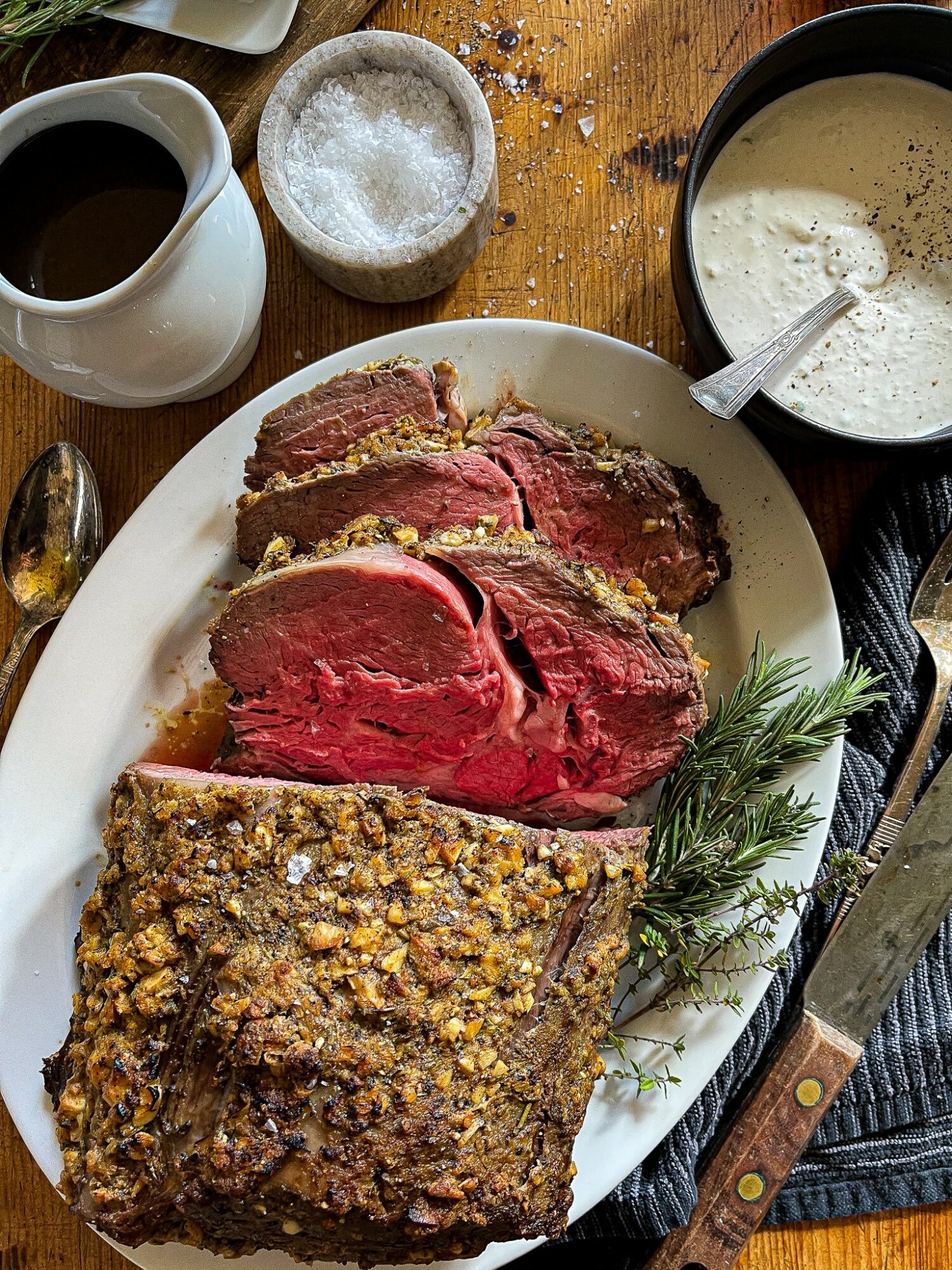 Cooked beef roast with a crust, sliced to show pink interior, garnished with herbs, on a white plate with sauces and condiments.