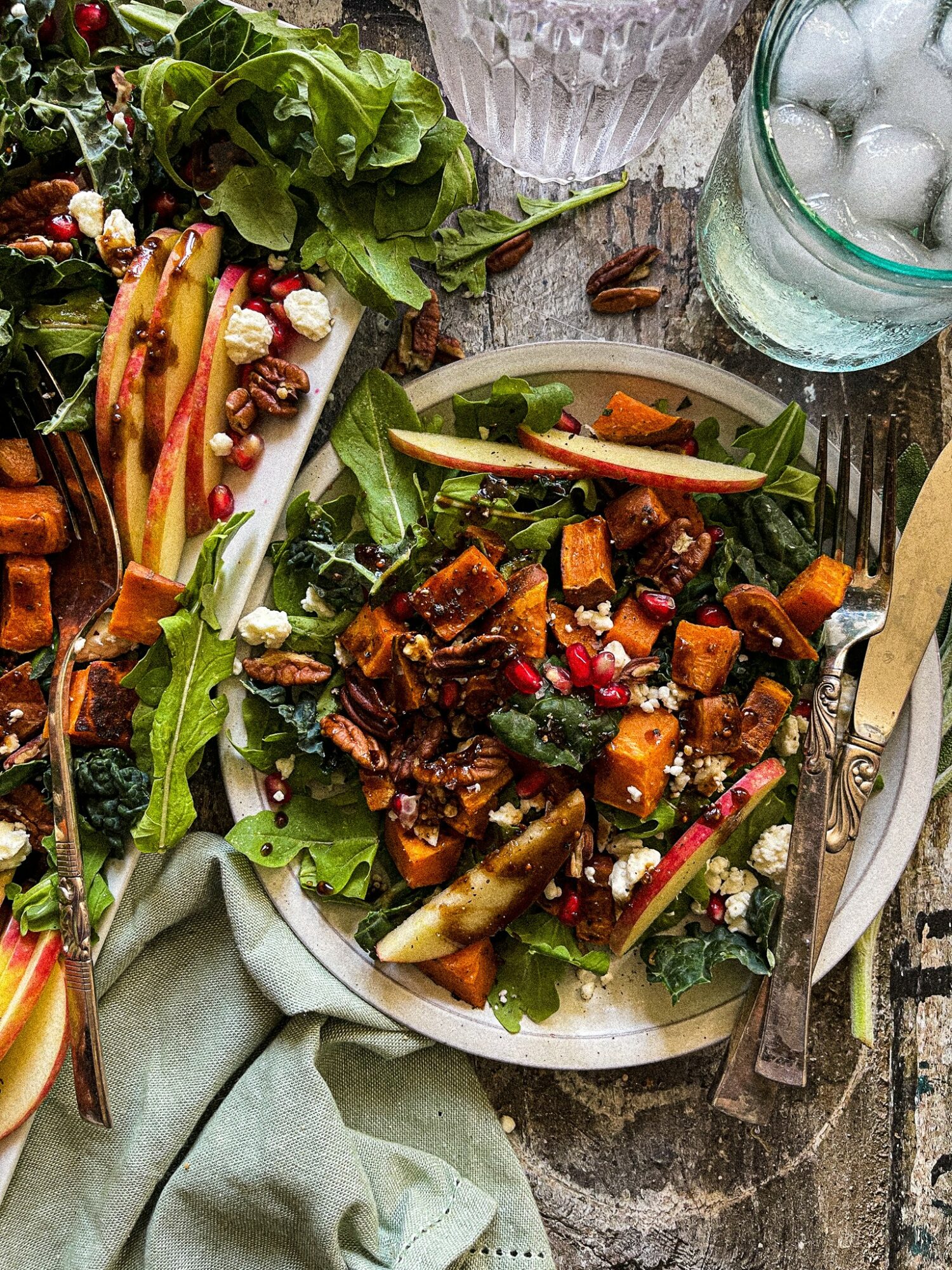 Salad with roasted vegetables, greens, and pomegranate seeds on a plate, with a glass of water and ice nearby.