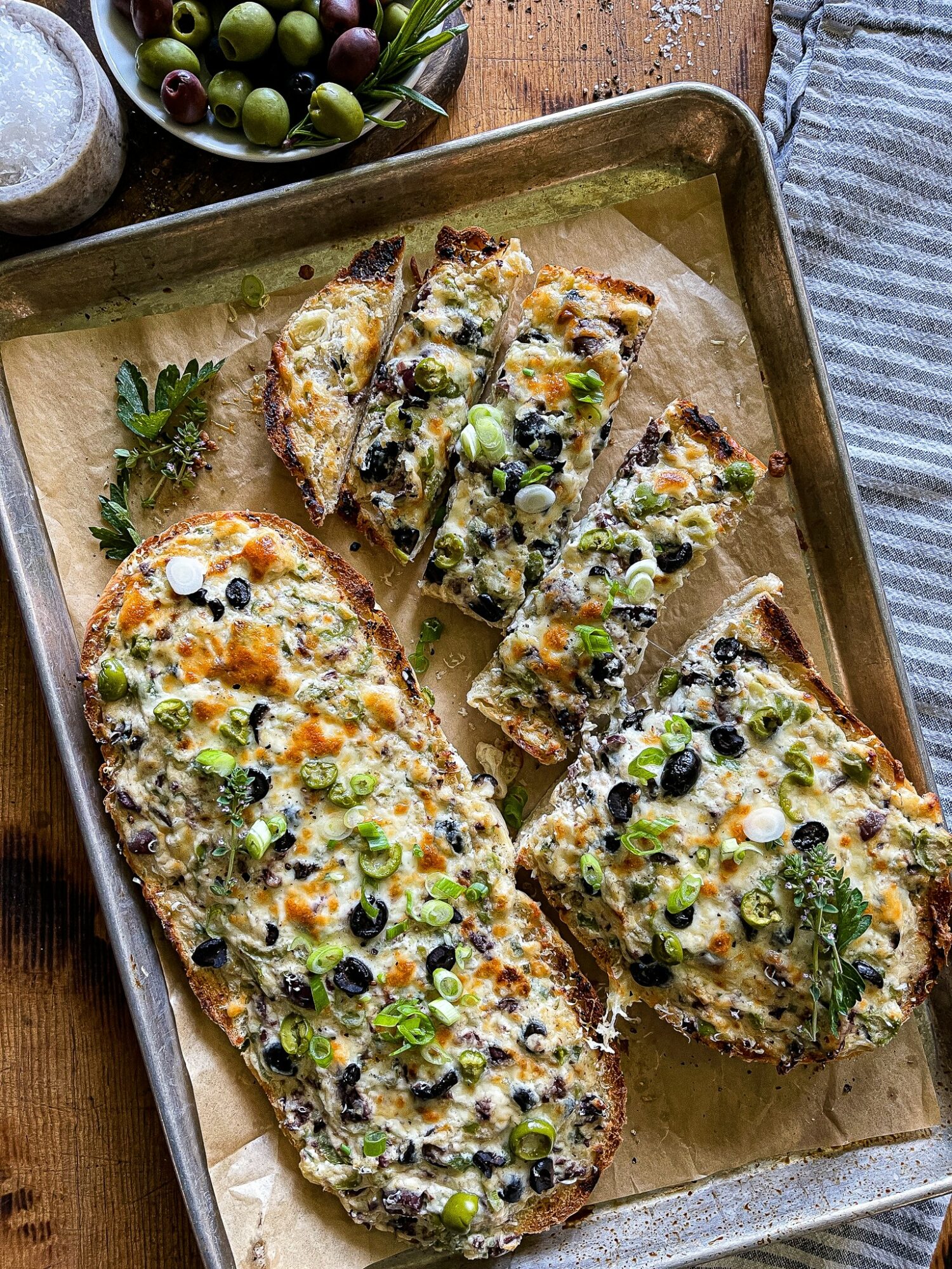 Four slices of vegetable and cheese bread on a baking sheet, garnished with herbs, with olives and grapes nearby.