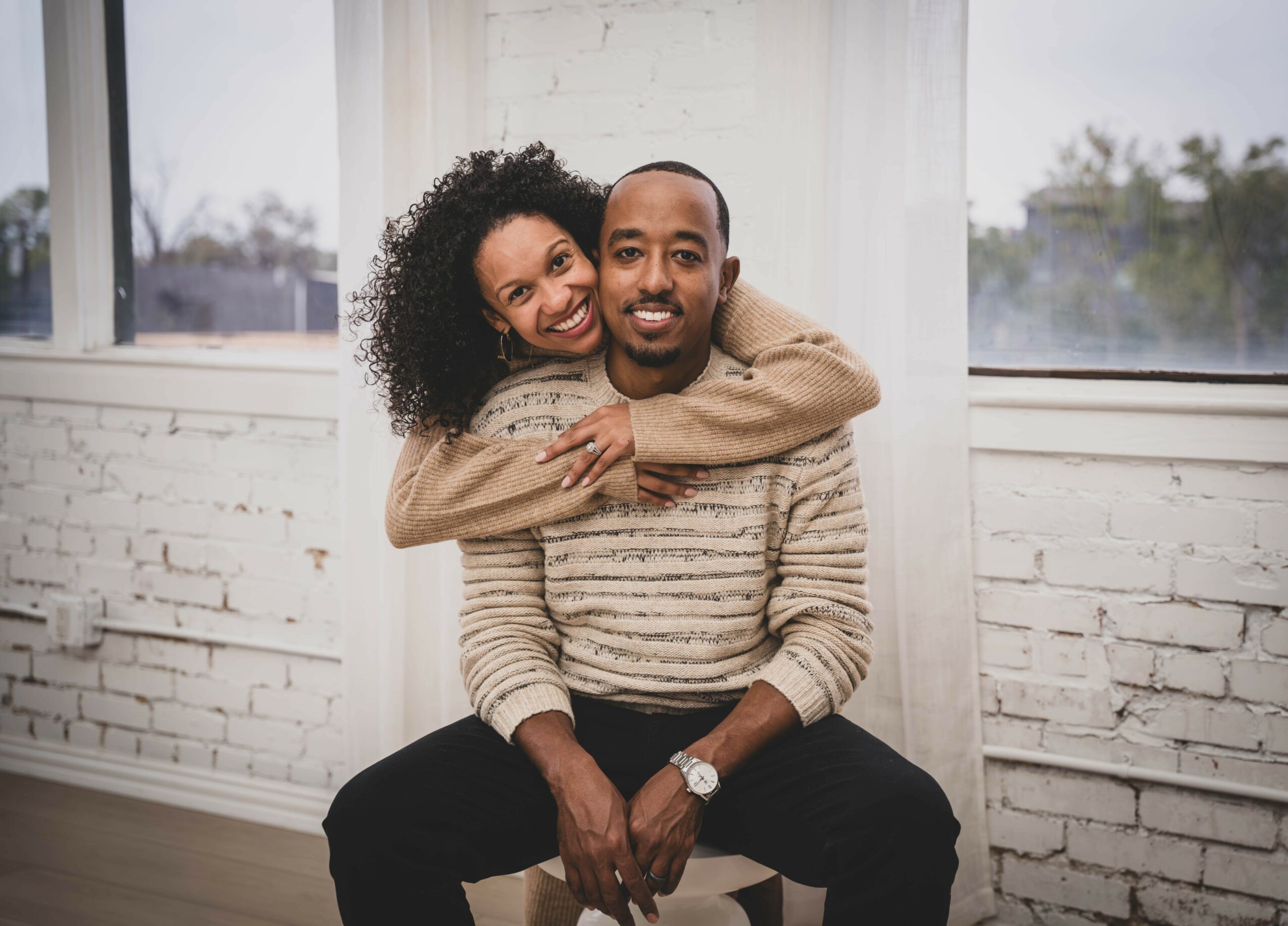A woman with curly hair hugs a man from behind, both smiling, sitting in a bright room with white brick walls.