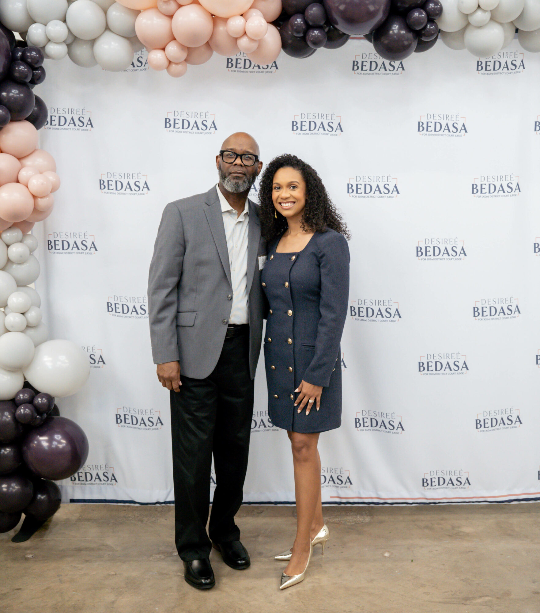 Man and woman standing together in front of a backdrop with balloons and 'BEDASA' text, smiling, dressed professionally.