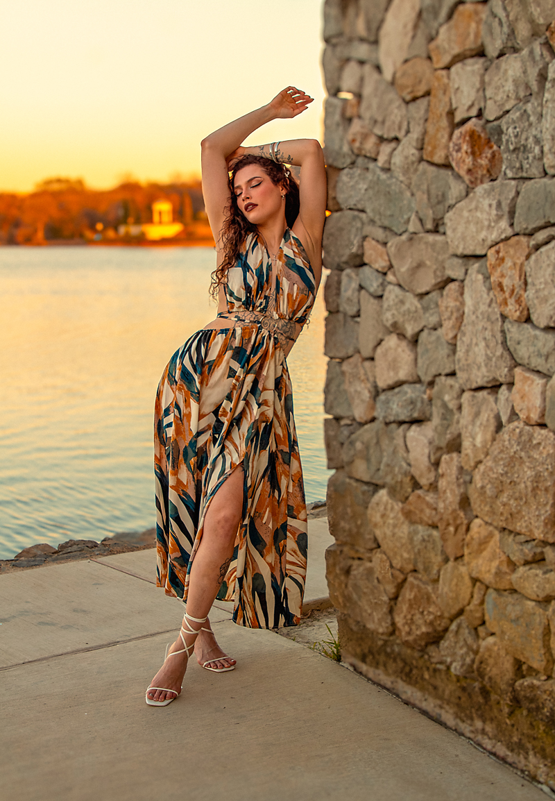 Woman in a patterned dress standing next to a stone wall near water at sunset, with arms raised above head.
