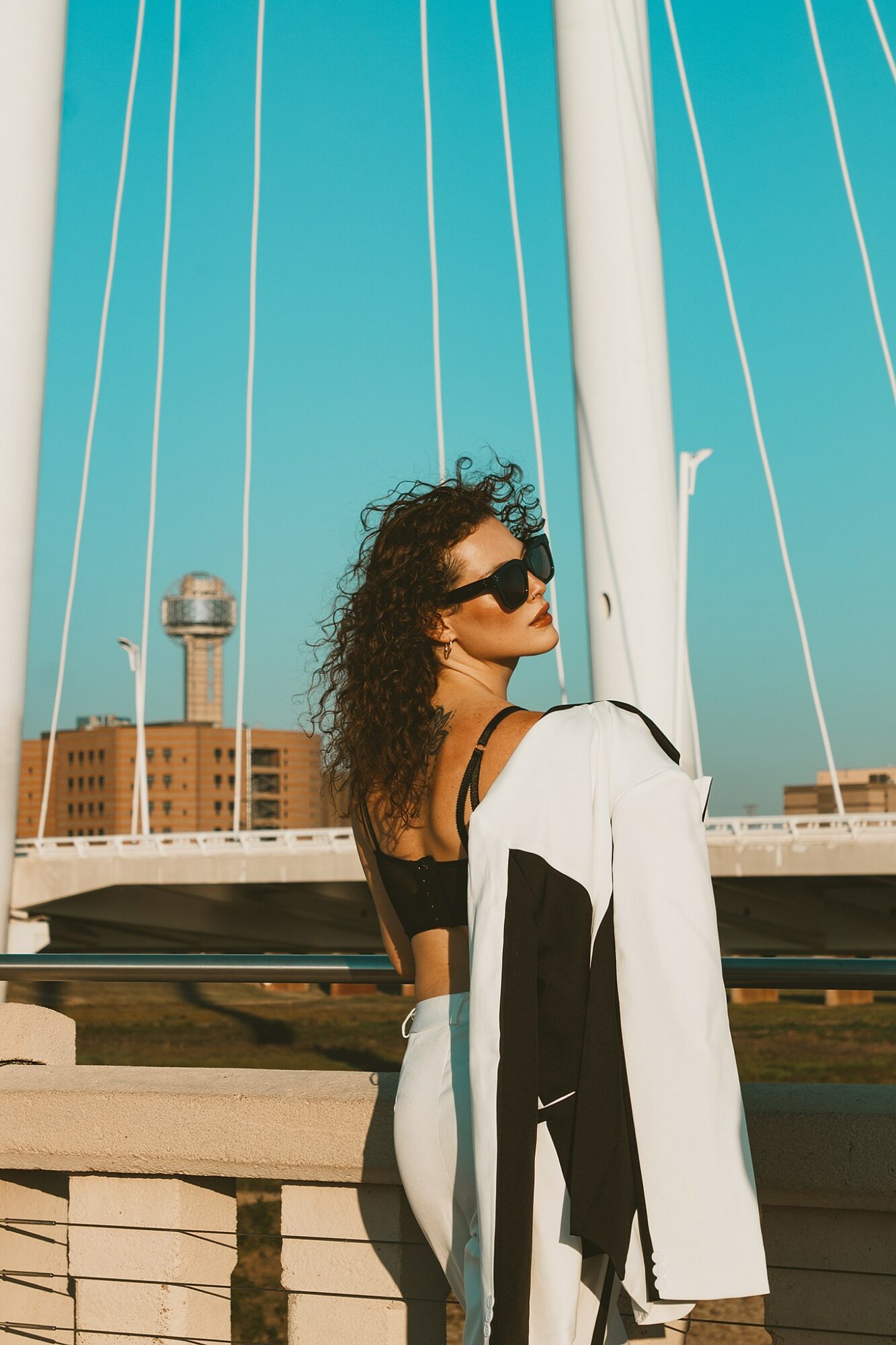 Woman with curly hair wearing sunglasses and a black top, standing near a marina with sailboats and buildings in background.