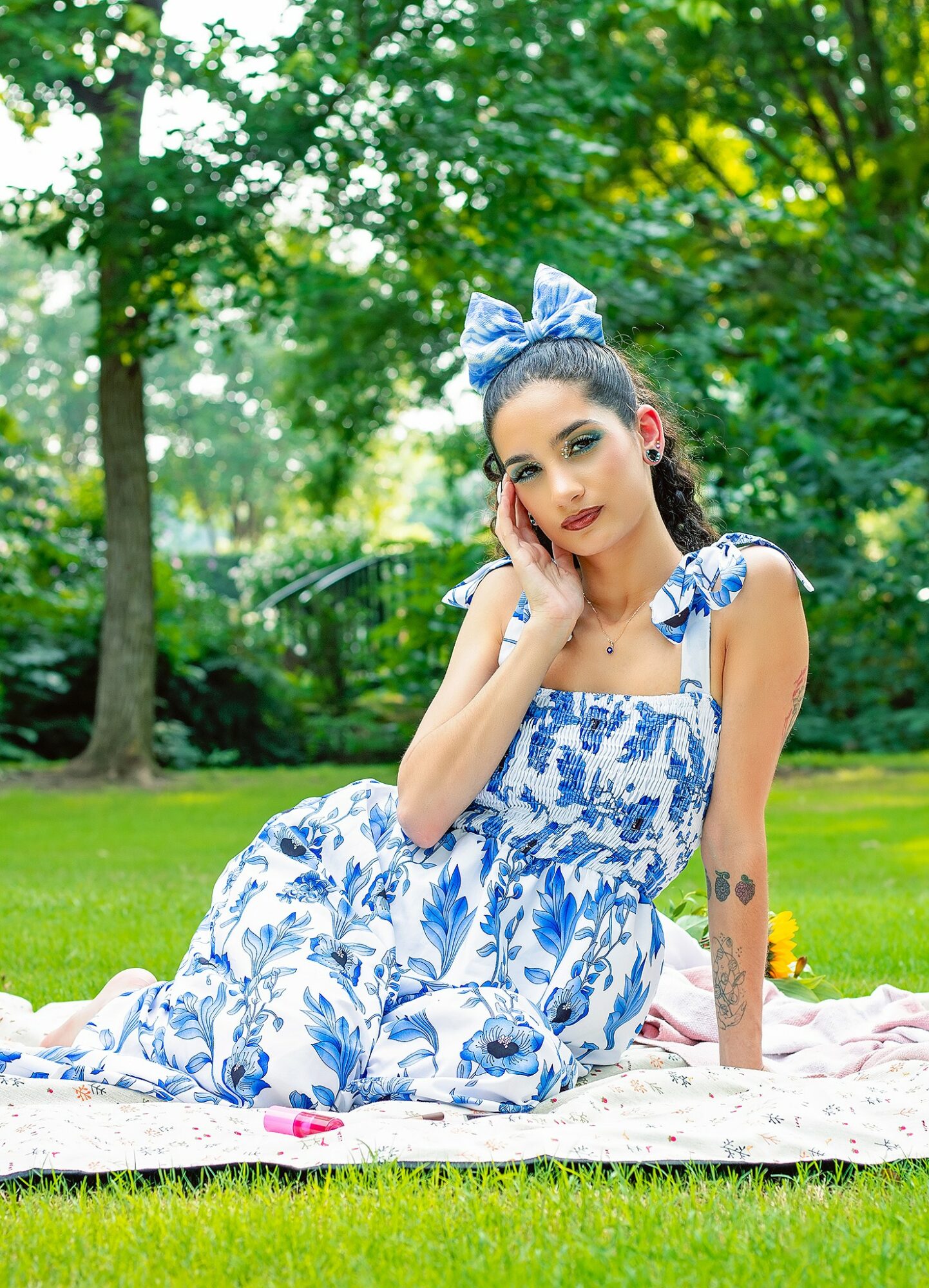 Young woman in a blue and white floral dress with a matching bow headband sitting on a blanket in a park with green trees.