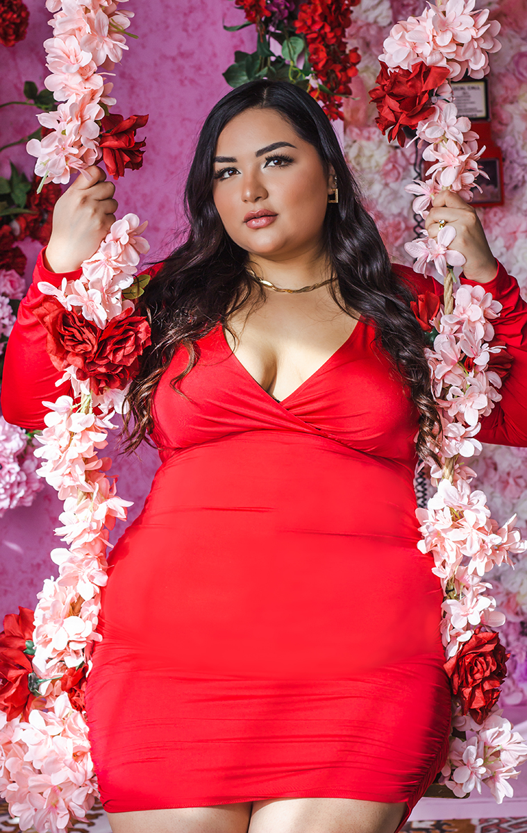 Woman in red dress standing among pink and red flowers, holding floral garlands, with a pink background.