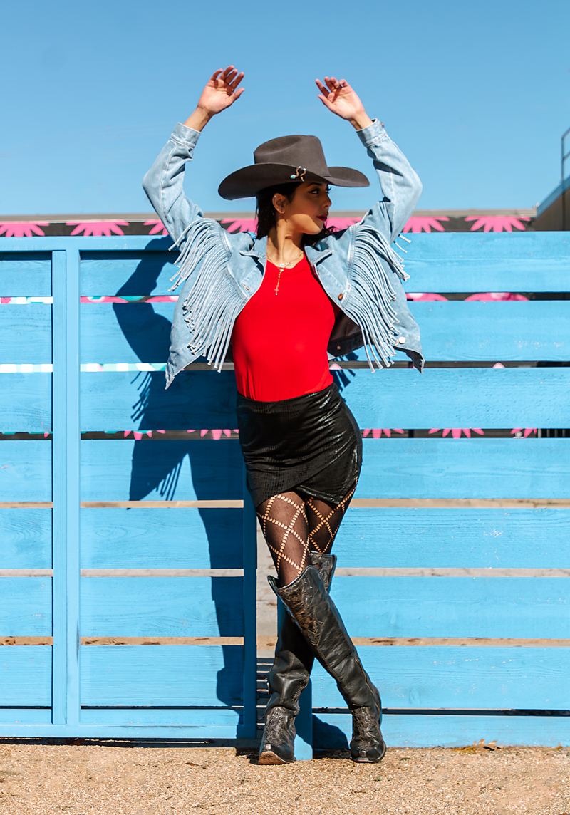 Woman in red top, black skirt, and tall boots stands against a blue fence with arms raised, wearing a wide-brimmed hat.