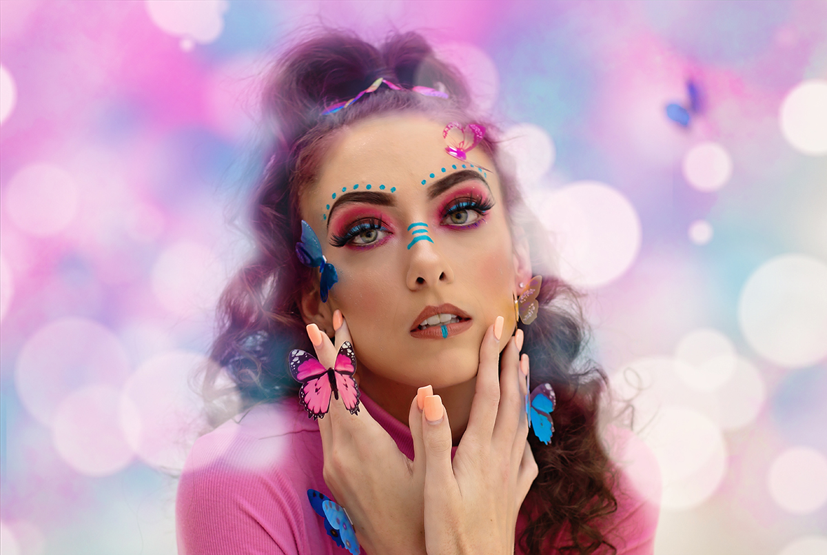 Woman with colorful makeup and butterfly accessories touches her face against a pink and purple background.