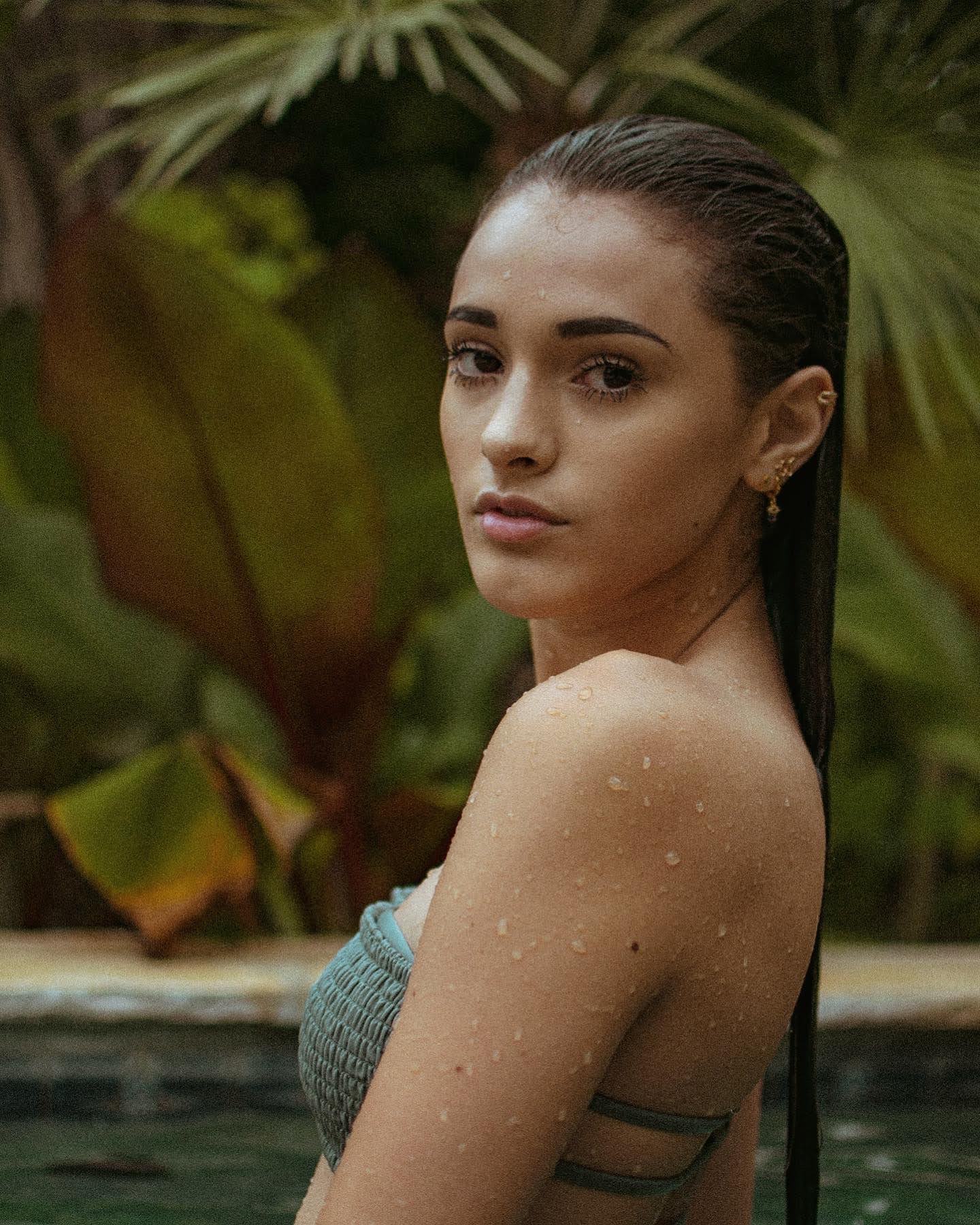 Young woman with wet hair and water droplets on her shoulder, looking at the camera outdoors with green foliage background.