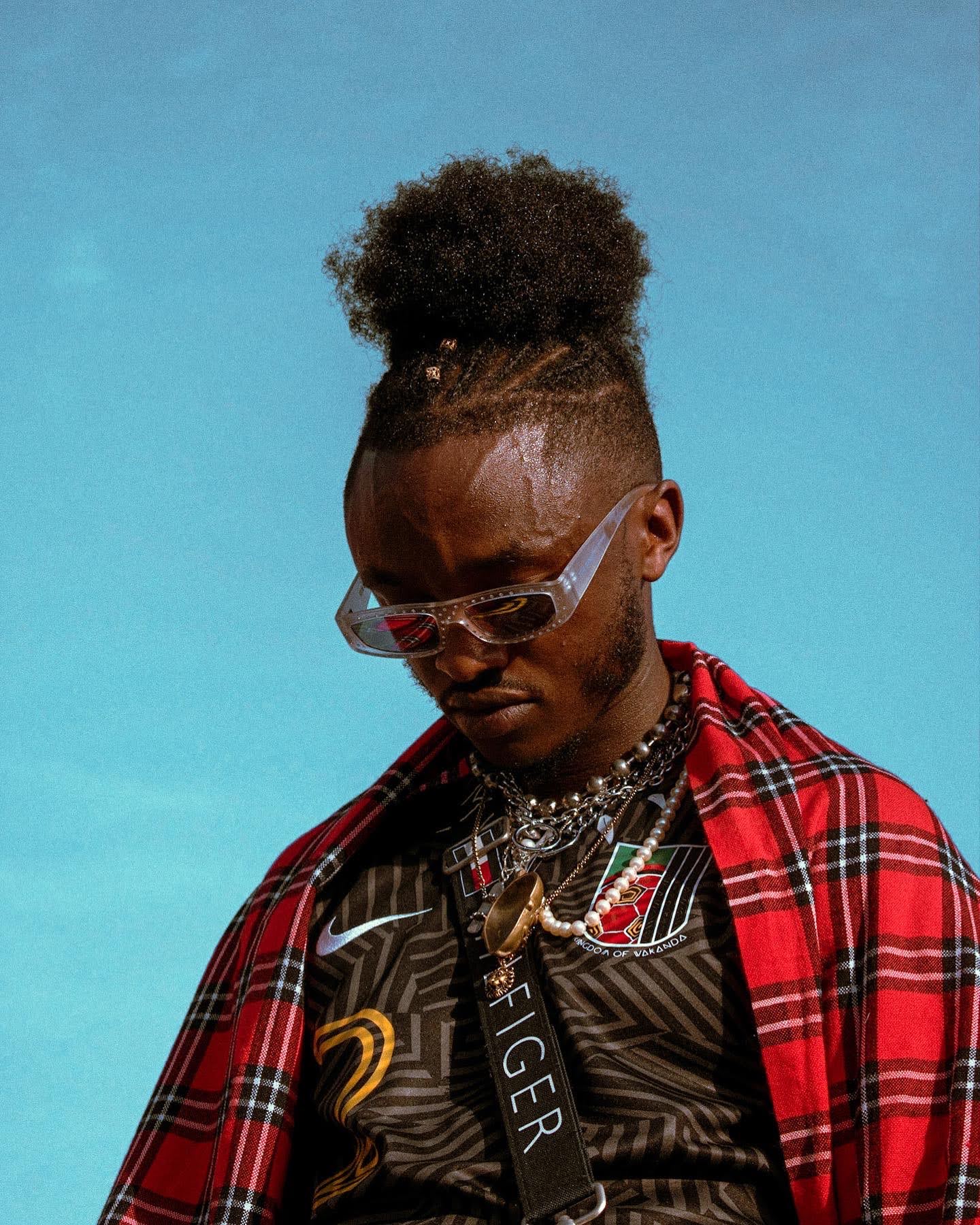 Person with curly hair in a high puff, wearing sunglasses, layered necklaces, a black sports jersey, and a red plaid shirt, against a blue background.