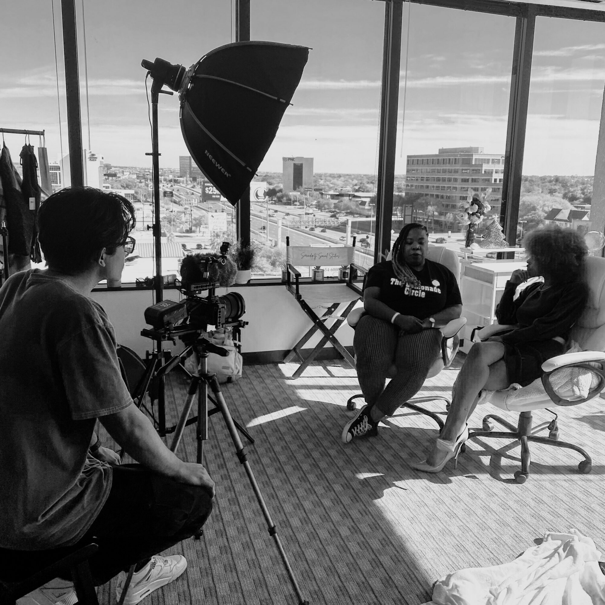 Two women sit in chairs during an interview in a room with large windows showing a cityscape, camera and lighting equipment present.