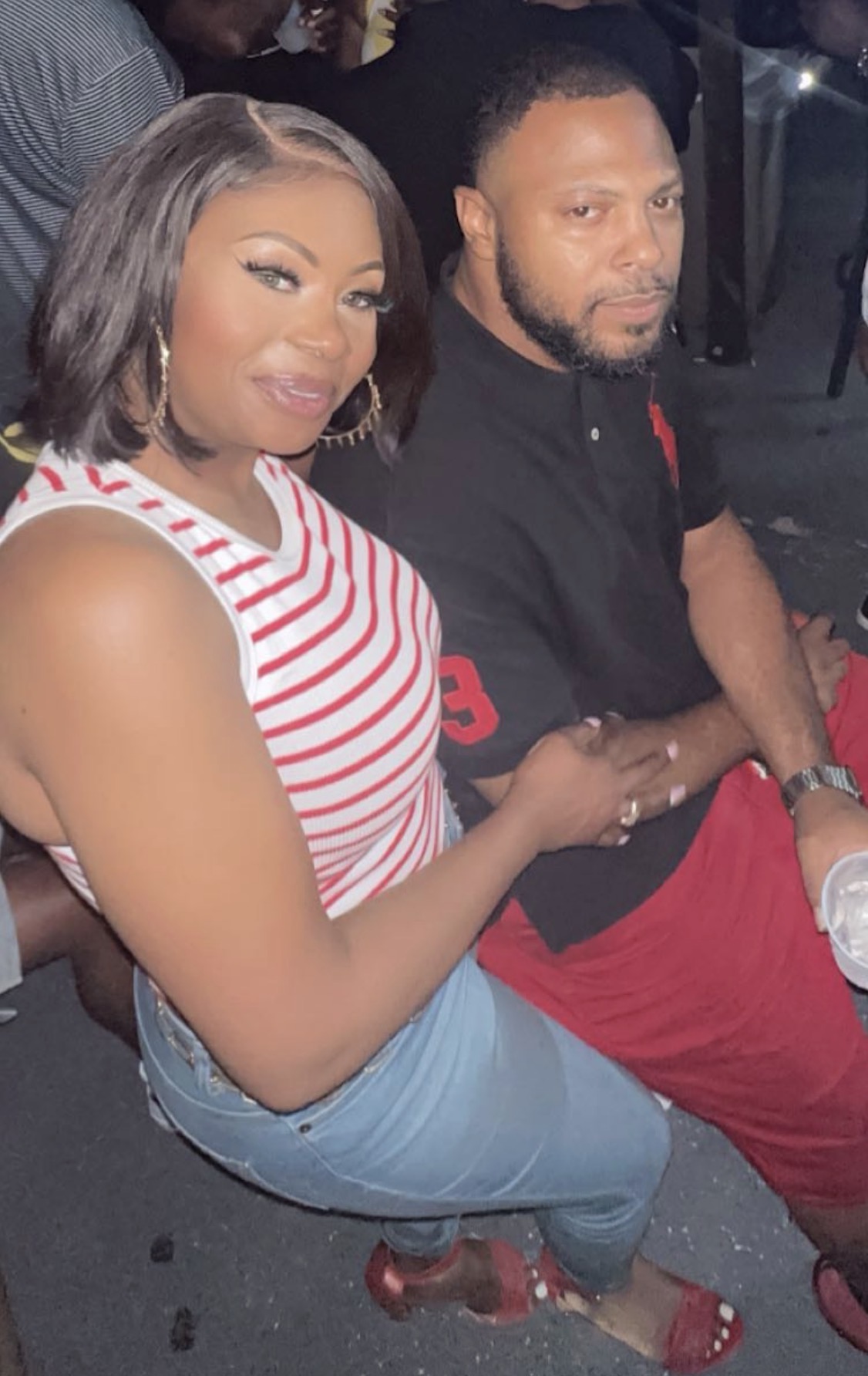 A woman and man sitting together, woman smiling, woman wearing a striped top and jeans, man in black shirt and red pants.