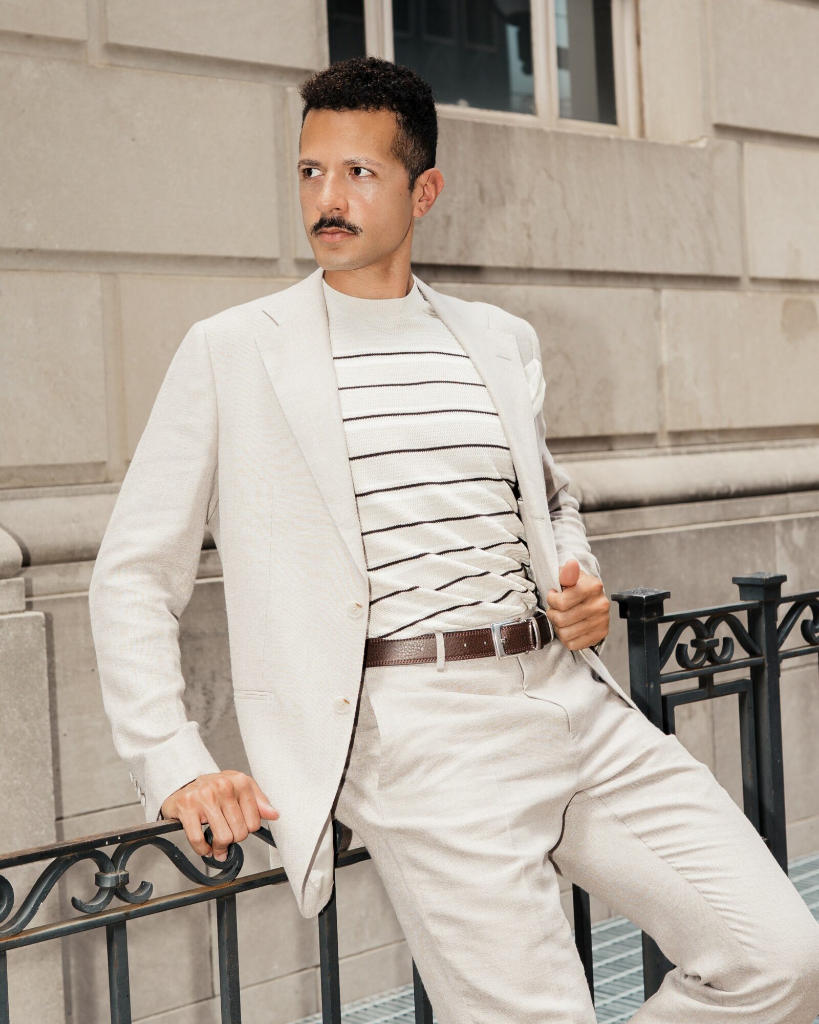 Man with a mustache in a white blazer and striped shirt leaning against a black railing outside a building.