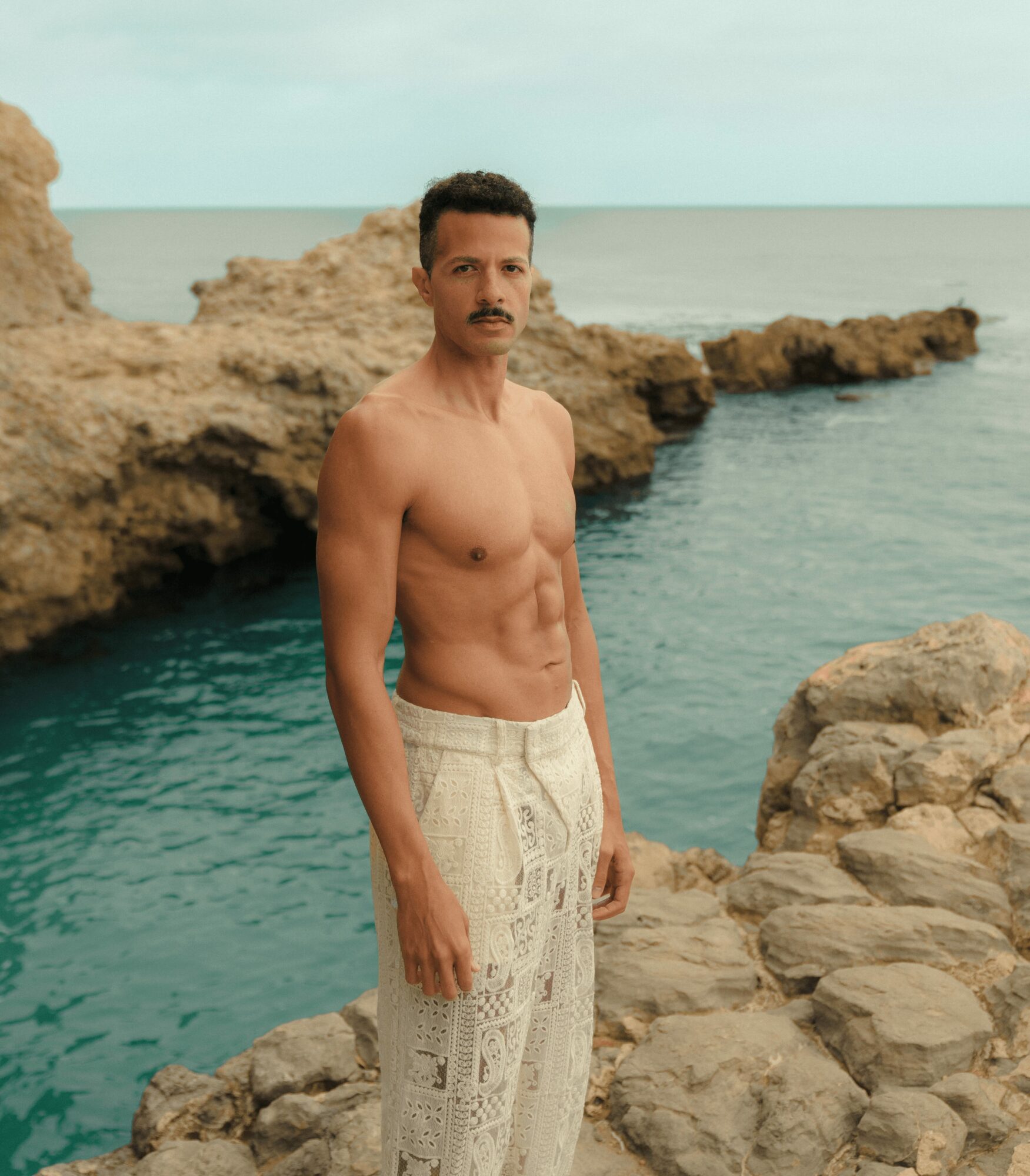 Man with a mustache and short dark hair stands shirtless on rocks by the water, wearing light-colored pants, with a rocky shoreline and ocean in background.