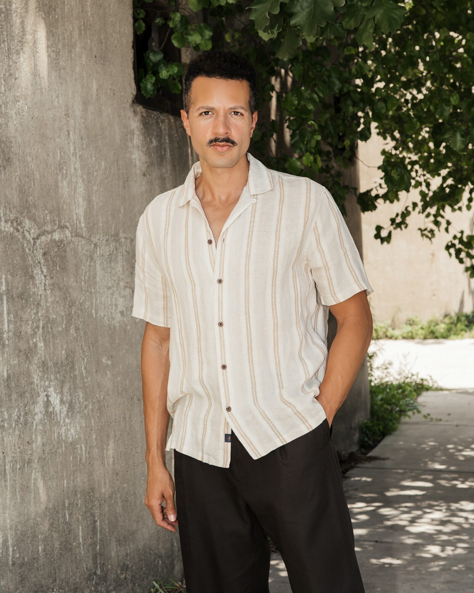 Man with dark hair and mustache wearing a striped short-sleeve shirt and dark pants, standing outdoors near a wall and trees.