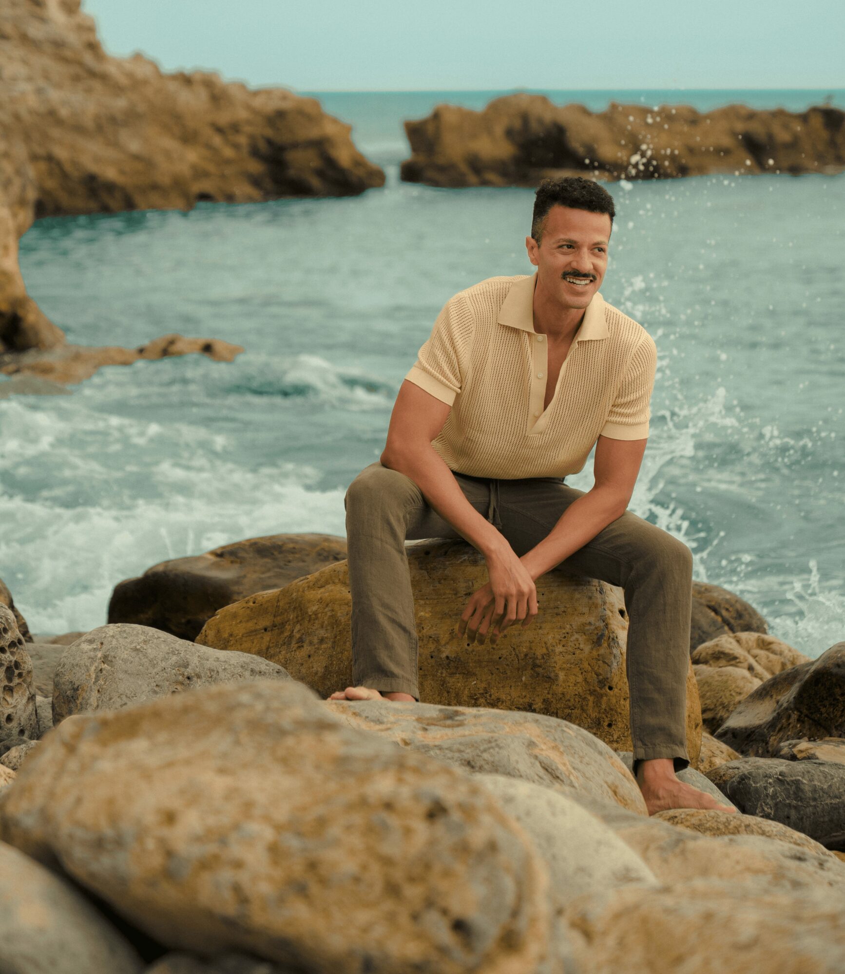 Man sitting on rocks by the ocean, smiling, with waves and rocky cliffs in background.