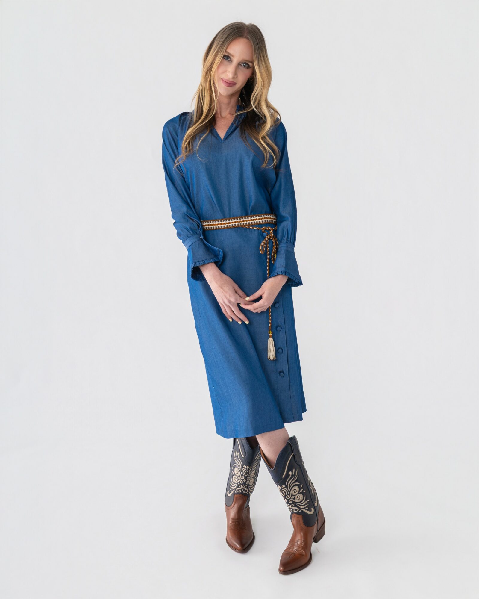 Woman in blue dress with a belt, standing with hands clasped, wearing cowboy boots, against a plain white background.